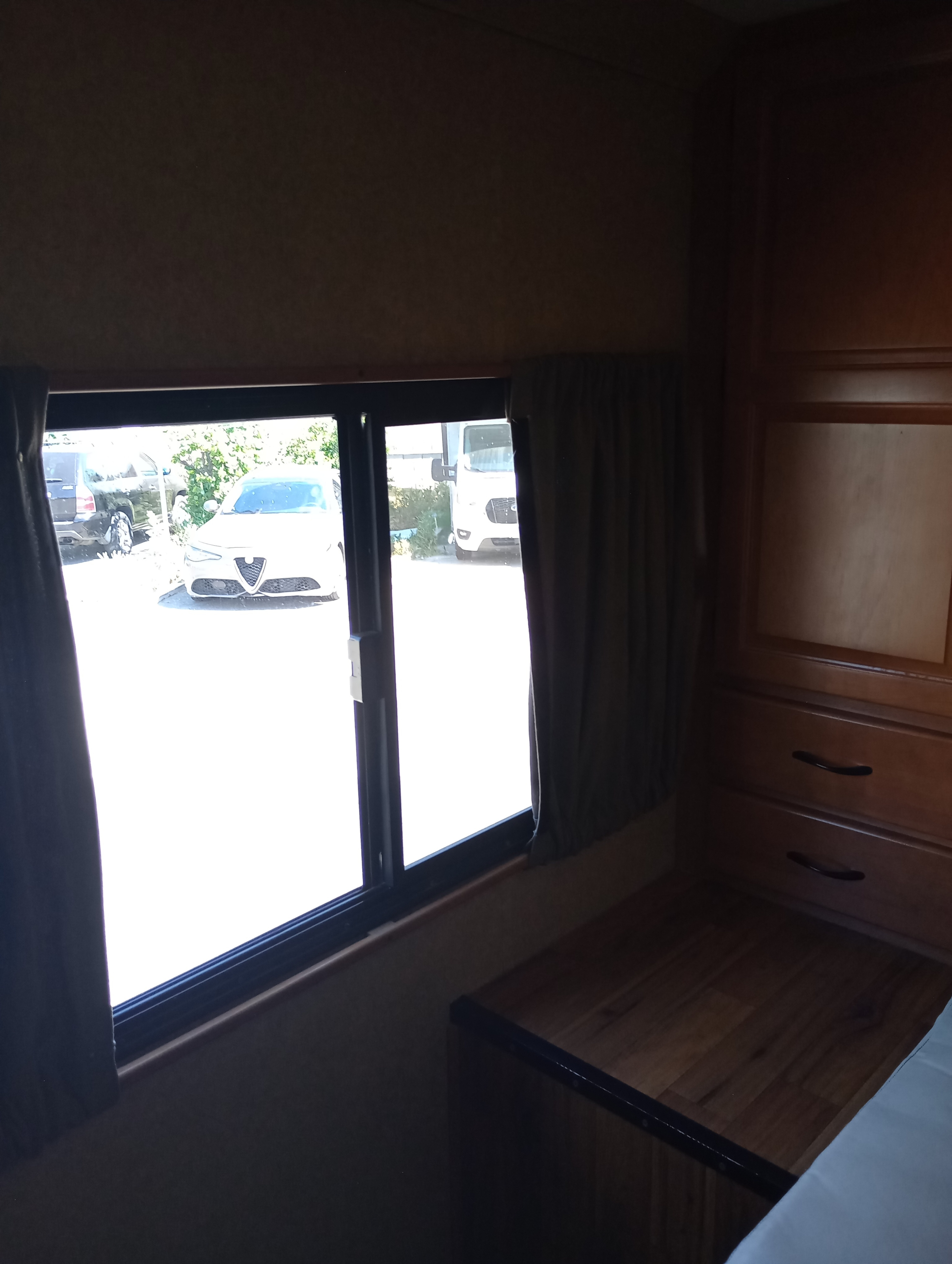 View from an RV rental's interior: a bright window shows parked cars, with dark curtains, wood cabinets, and a bed inside.. Ford Majestic 2019