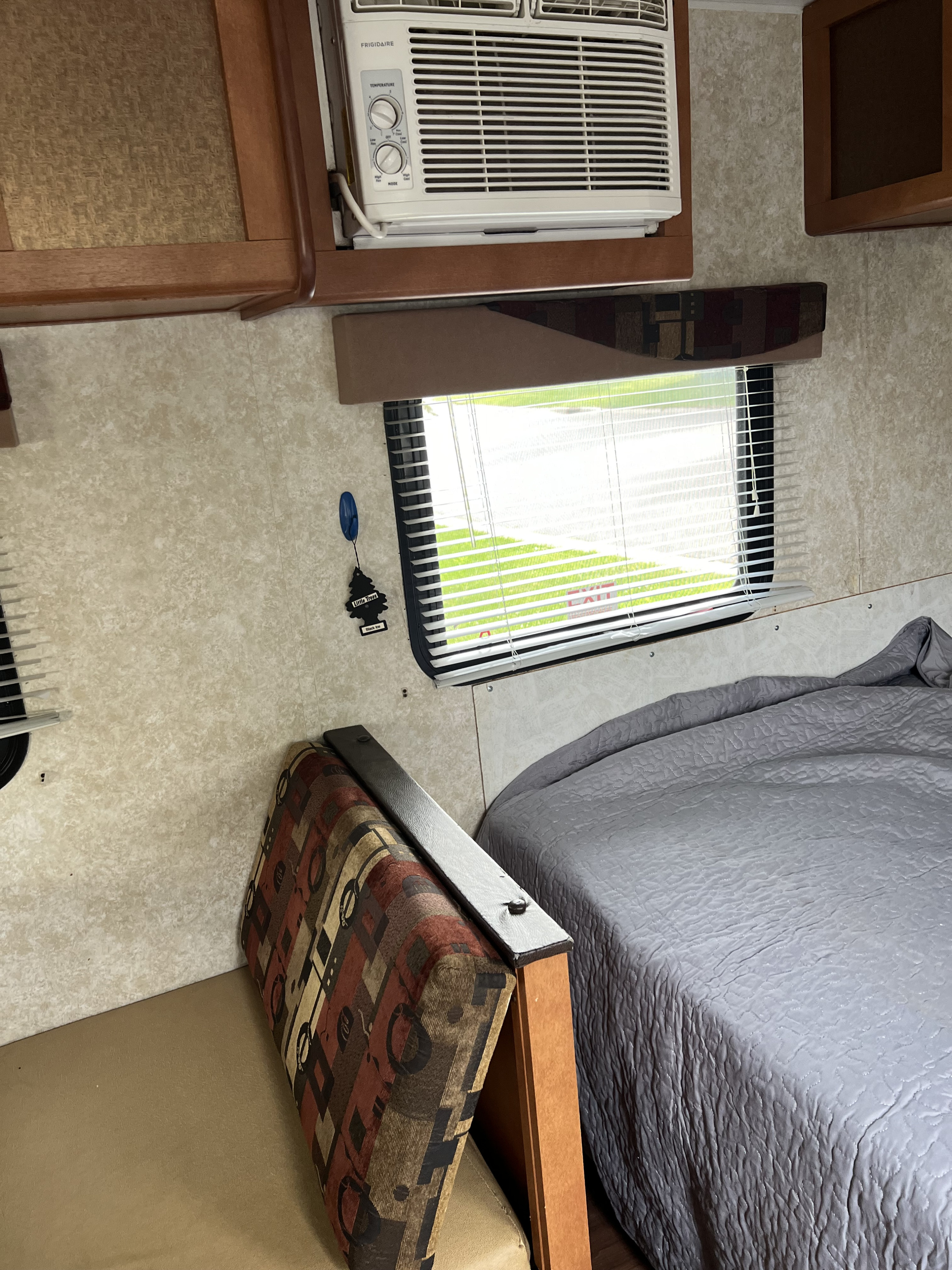 Interior of an rv rental: features a bed with grey comforter, a patterned dinette seat, wall-mounted AC unit, and window with blinds.. Gulf Stream Ameri-Lite 2018