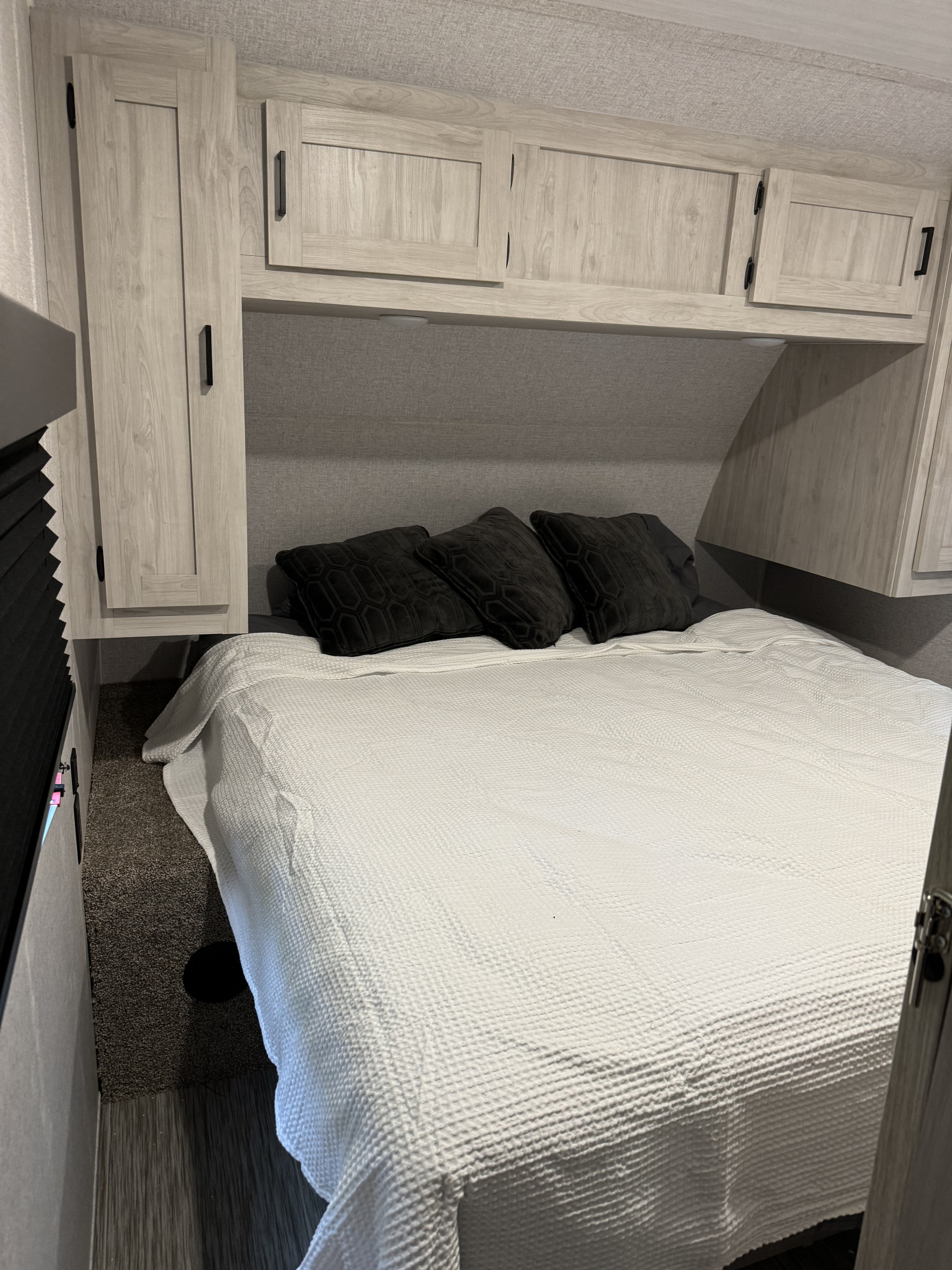 Inside an RV rental: a cozy bedroom featuring a white bedspread, dark pillows, and light wood-look cabinetry.. East to West Silver Lake 2023