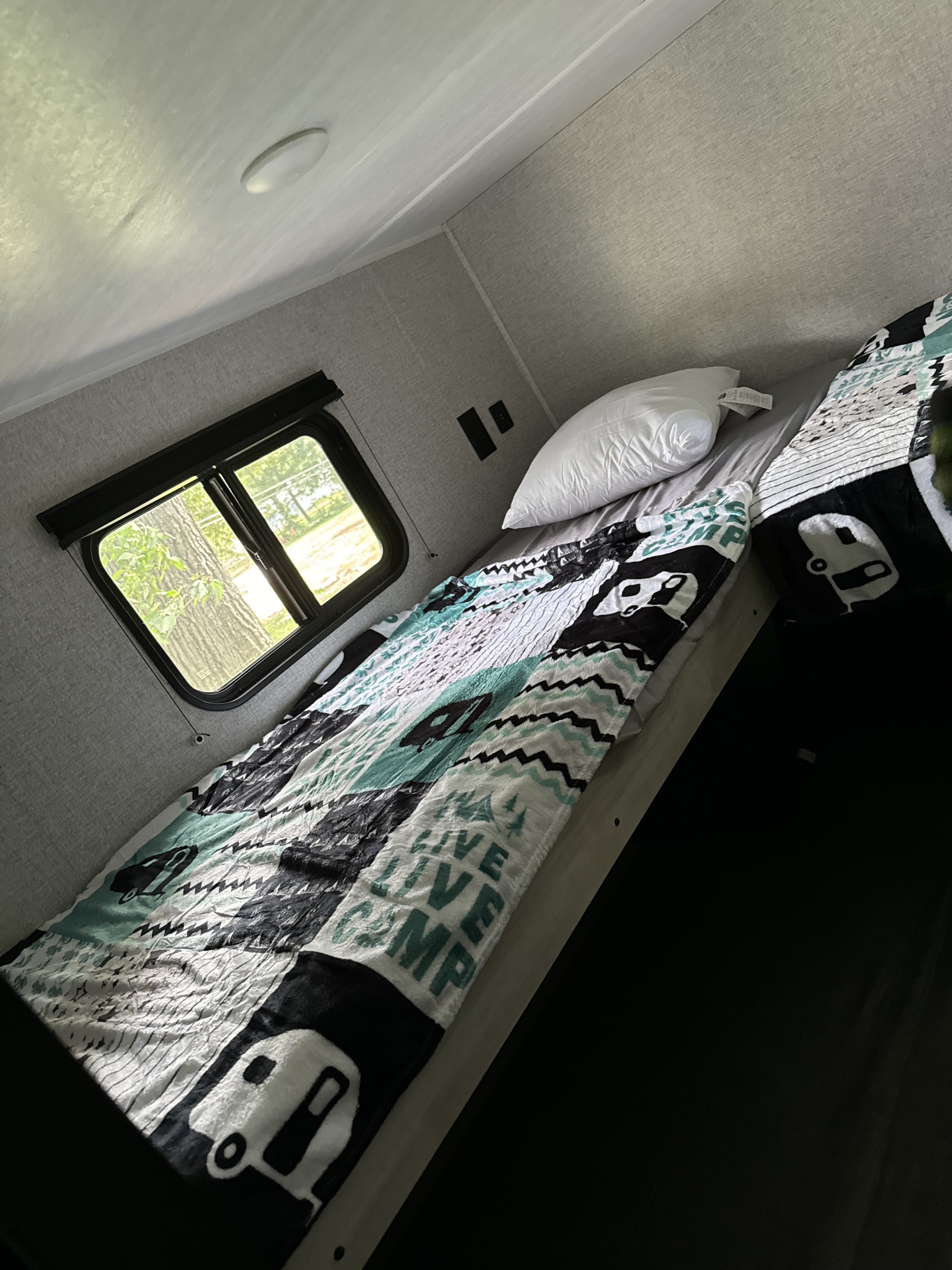 Comfortable RV rental bunk bed area with a camper-themed blanket, white pillow, and a bright window view of nature.. East to West Silver Lake 2023