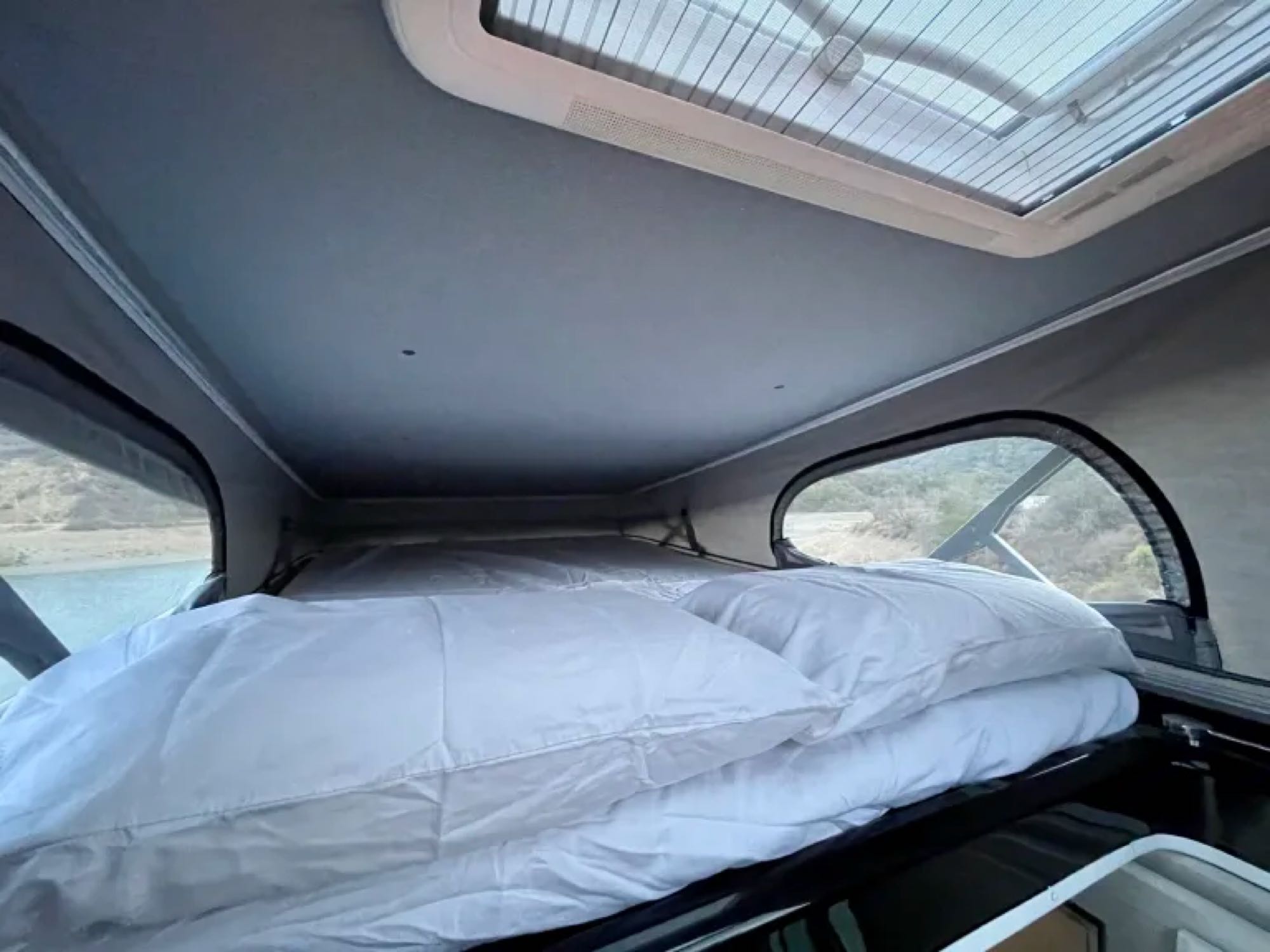 Experience comfort in this pop-top RV rental! A bed with white linens, large windows, and skylight offer scenic views.. Thor Tellaro 2024