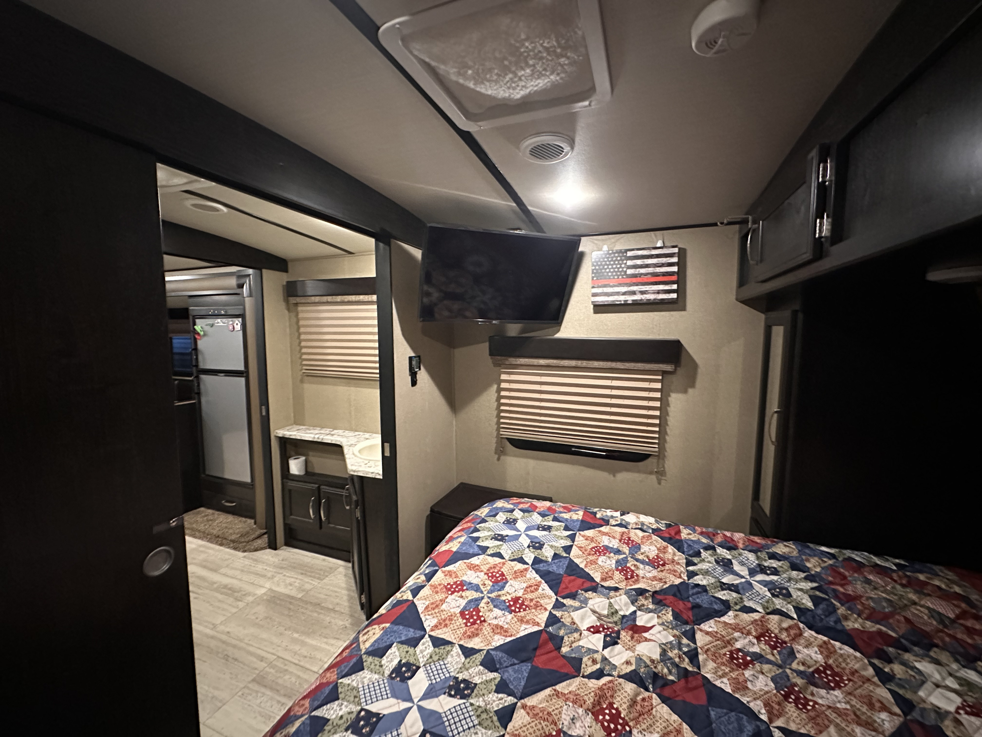 An RV rental's bedroom area, with a vibrant quilt, wall-mounted TV, and a view of the adjacent living space.. Grand Design Imagine 2018