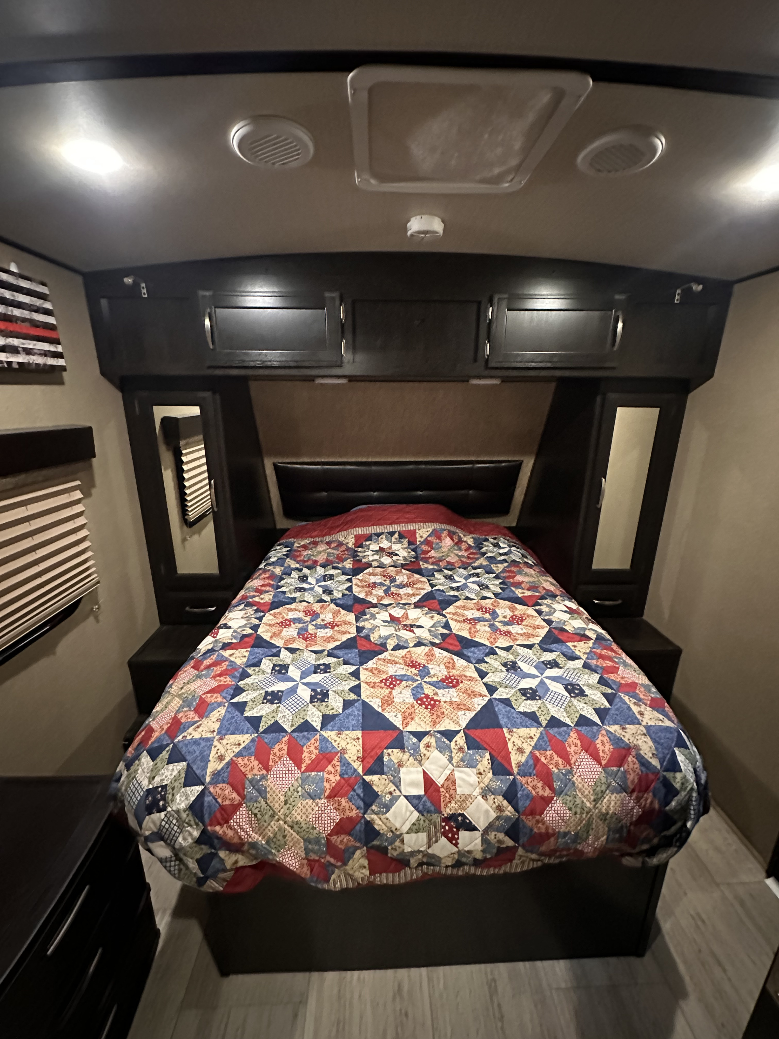 RV rental interior: A cozy bedroom featuring a bed with a colorful patchwork quilt, built-in dark wood storage, and overhead lighting.. Grand Design Imagine 2018