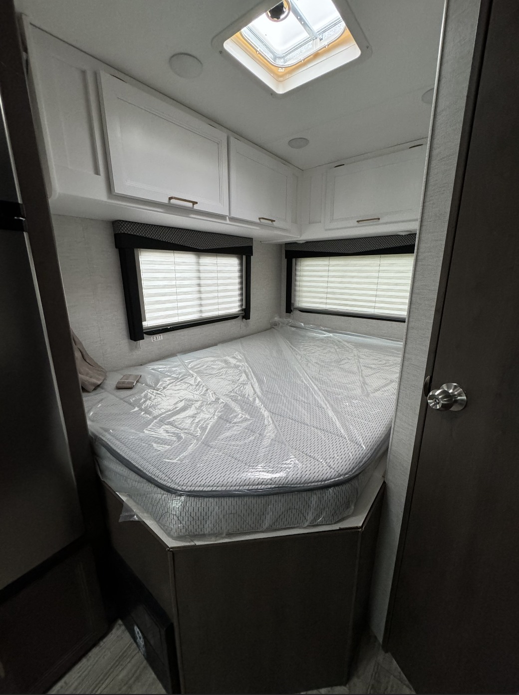 Bright RV rental bedroom features a new, plastic-covered mattress, white overhead cabinets, and dual windows with blinds.. Thor Motor Coach Freedom Elite 2024