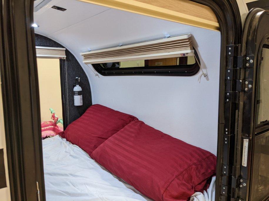 Cozy RV rental sleeping area with red pillows, white sheets, window blinds, and a mounted bottle inside.. Little Guy Little Guy Trailer 2018
