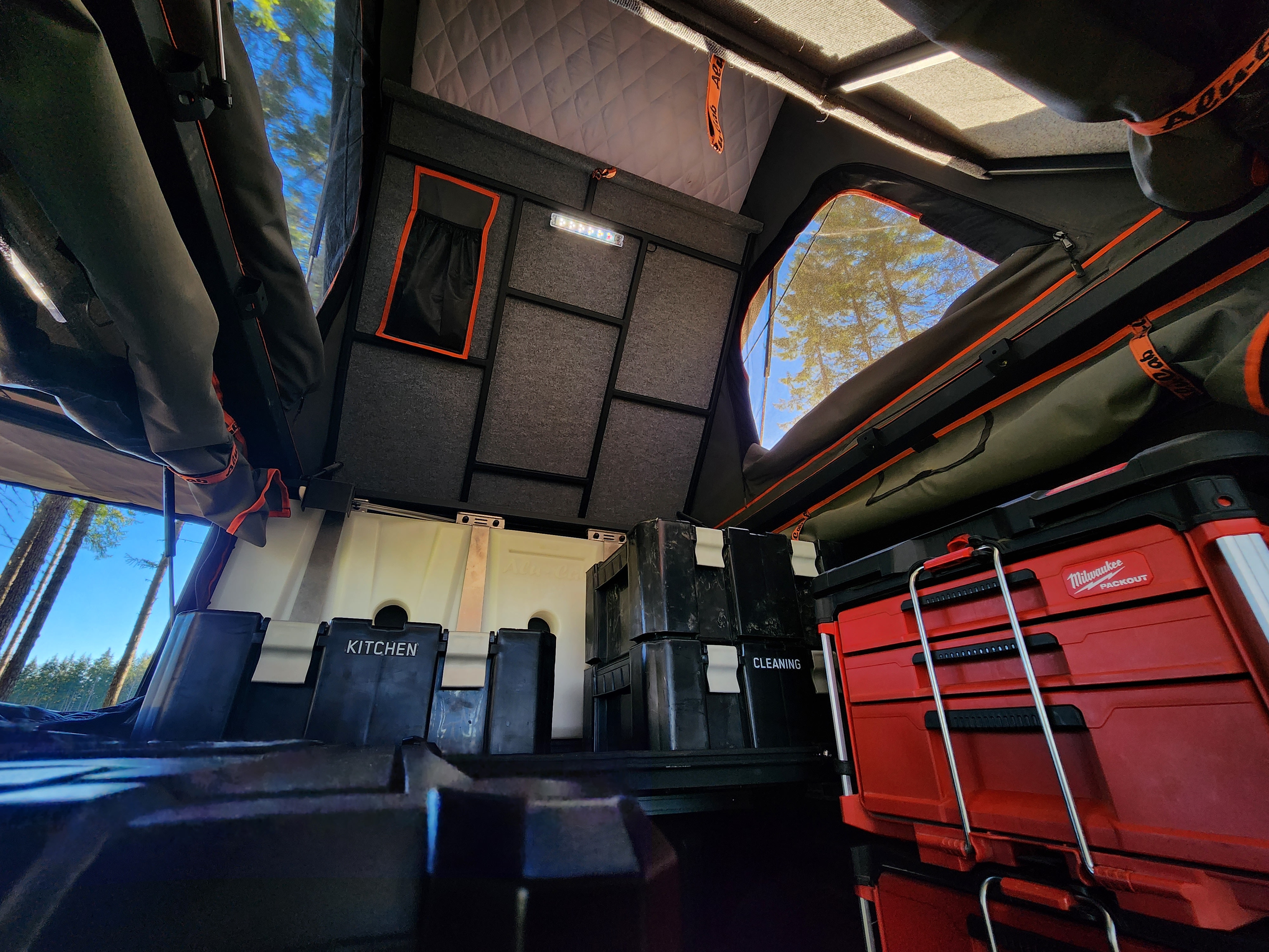 Inside a well-equipped truck camper, great for RV rental. Features organized storage like kitchen bins & Milwaukee Packout, with forest views.. Nissan Frontier 2022