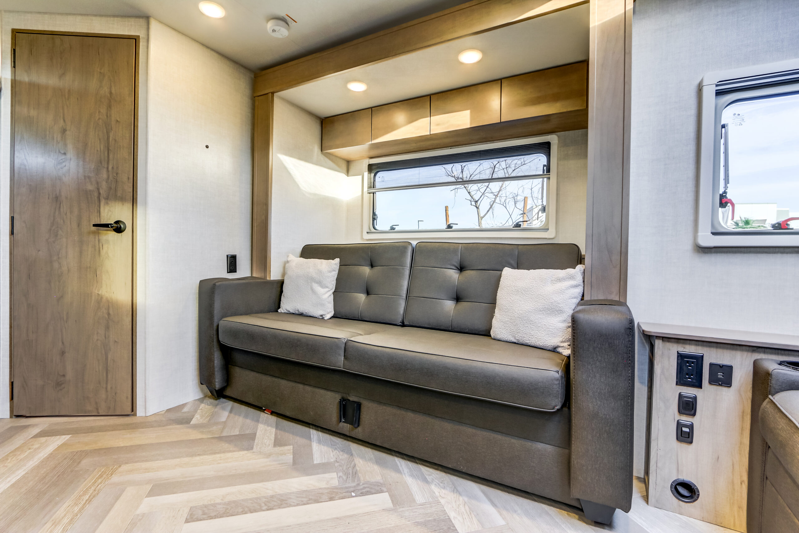Bright RV rental living space: gray sofa, large window, overhead cabinets, and wood herringbone floor.. Ember Overland 2023