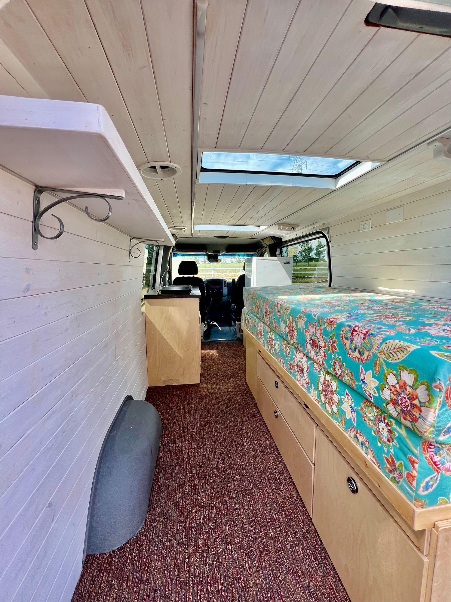 This RV rental interior features a cozy bed with floral cover, storage, skylight, and light wood walls for comfortable travel.. Mercedes Sprinter 2016