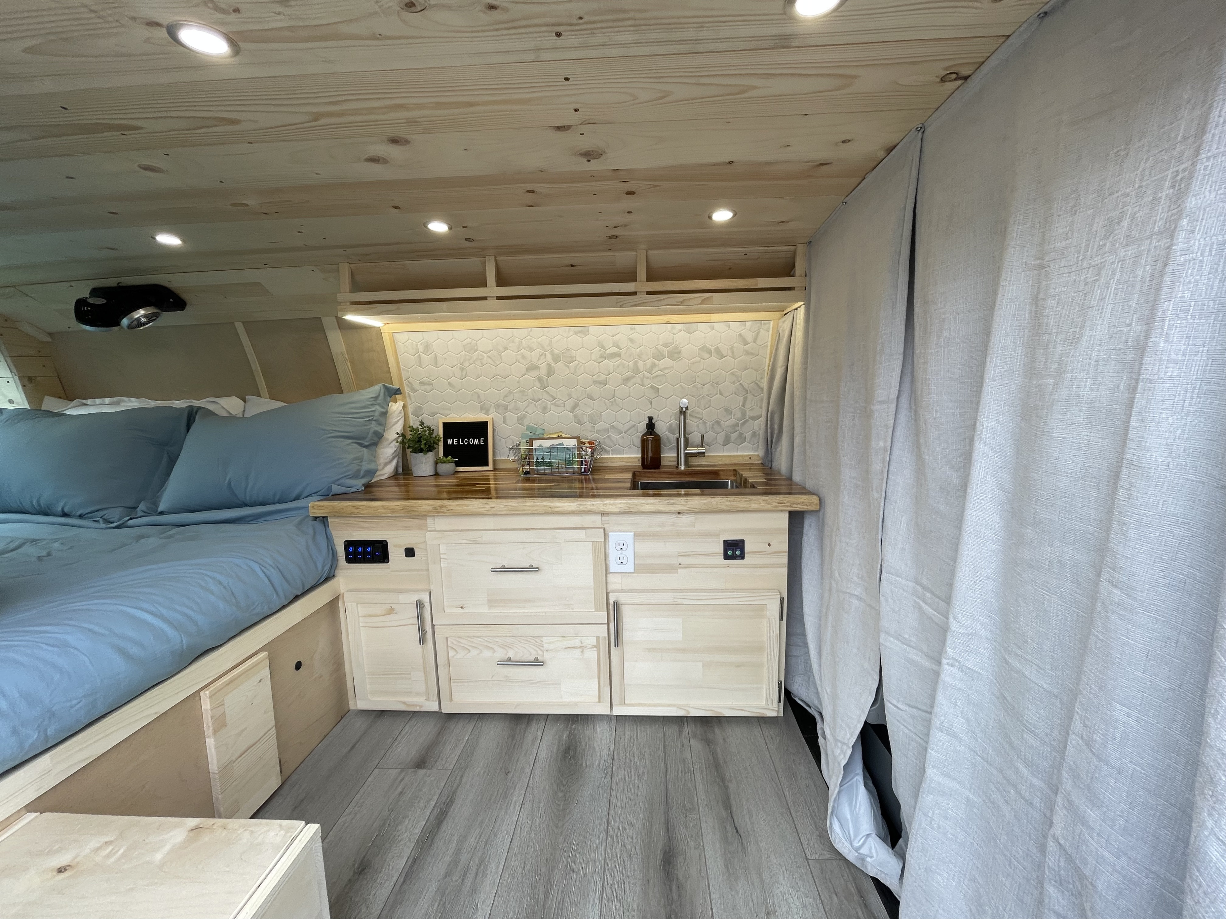 Bright RV rental interior: wood-paneled ceiling, blue bed on left, light wood kitchen with sink and "WELCOME" sign, grey curtain.. Gmc Savanna 2015