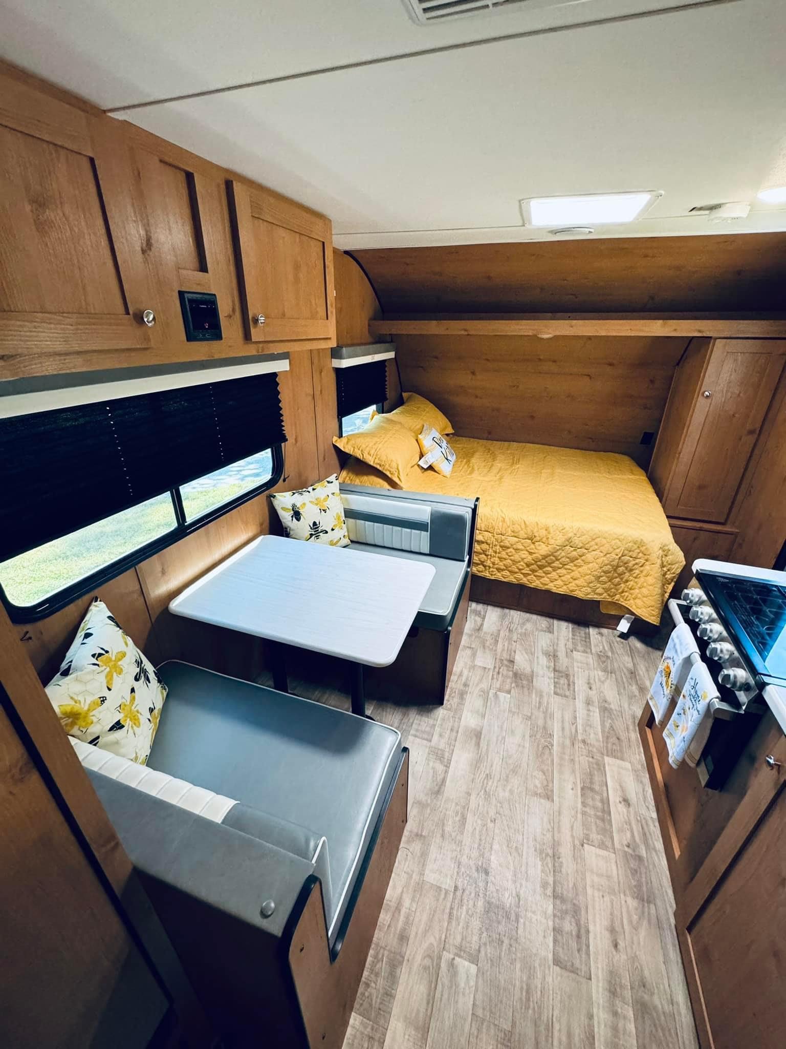 View of a compact RV rental interior featuring a dinette with grey cushions, bed with yellow quilt, and stove.. Riverside RV Retro 2023
