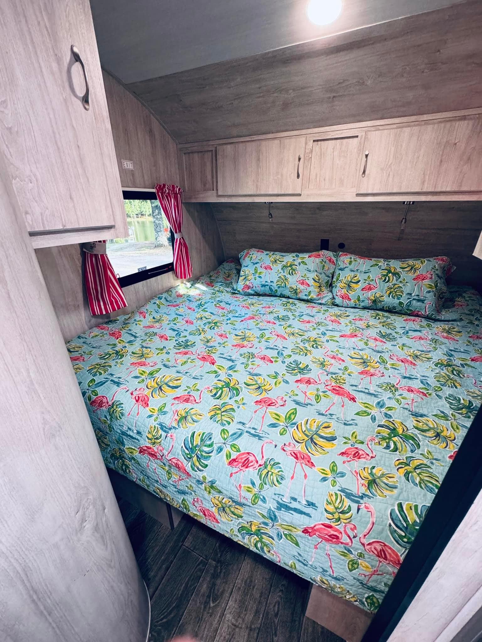 Inside an RV rental: a bright bedroom features a flamingo-patterned bed, wood-grain interior, and a lakeside window view.. Gulf Stream Vintage Cruiser 2024