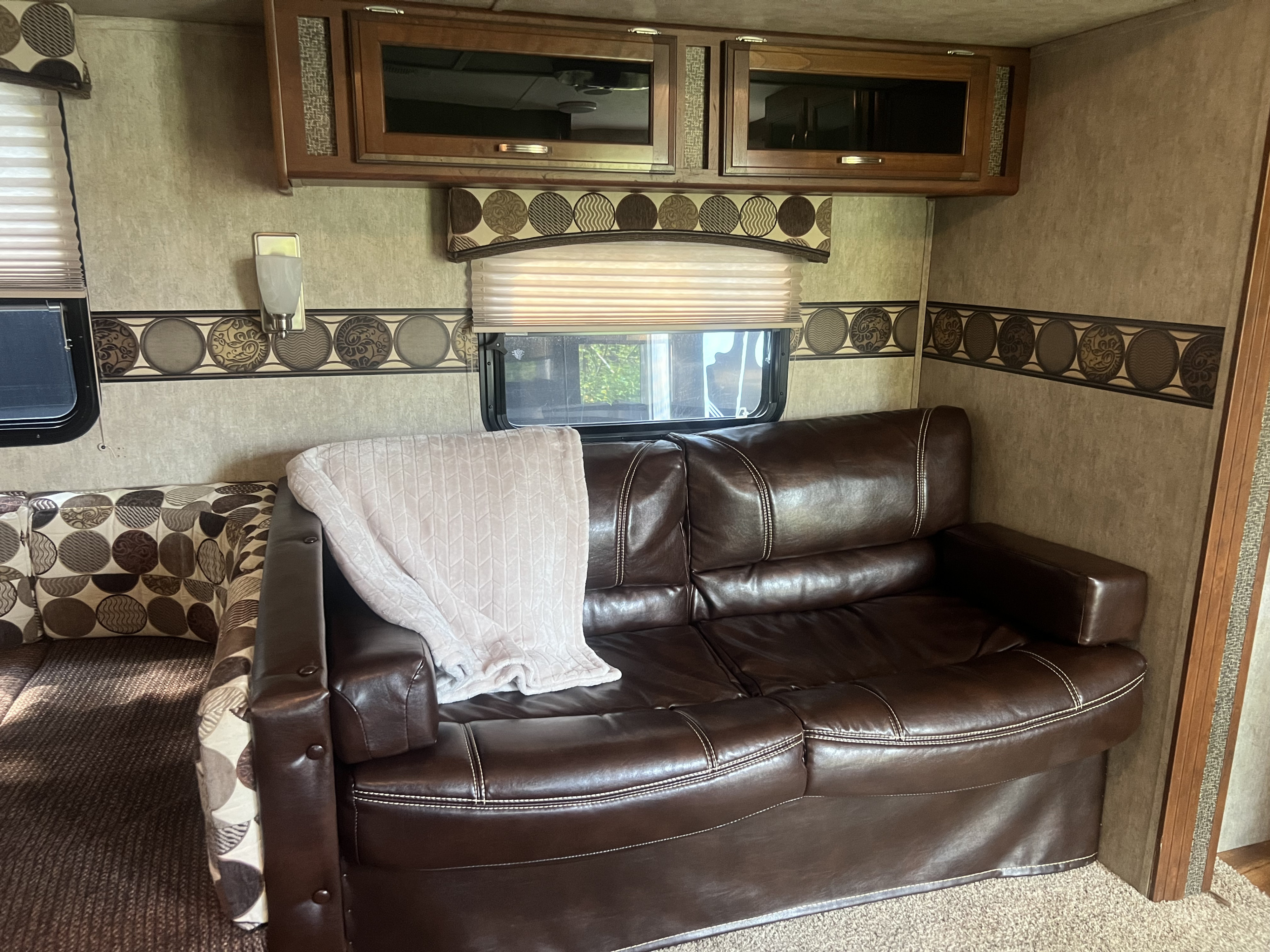 Cozy RV rental interior featuring a brown leather-like sofa, overhead cabinets, and patterned decor. Ideal for travel adventures.. Keystone RV Passport Grand Touring 2015