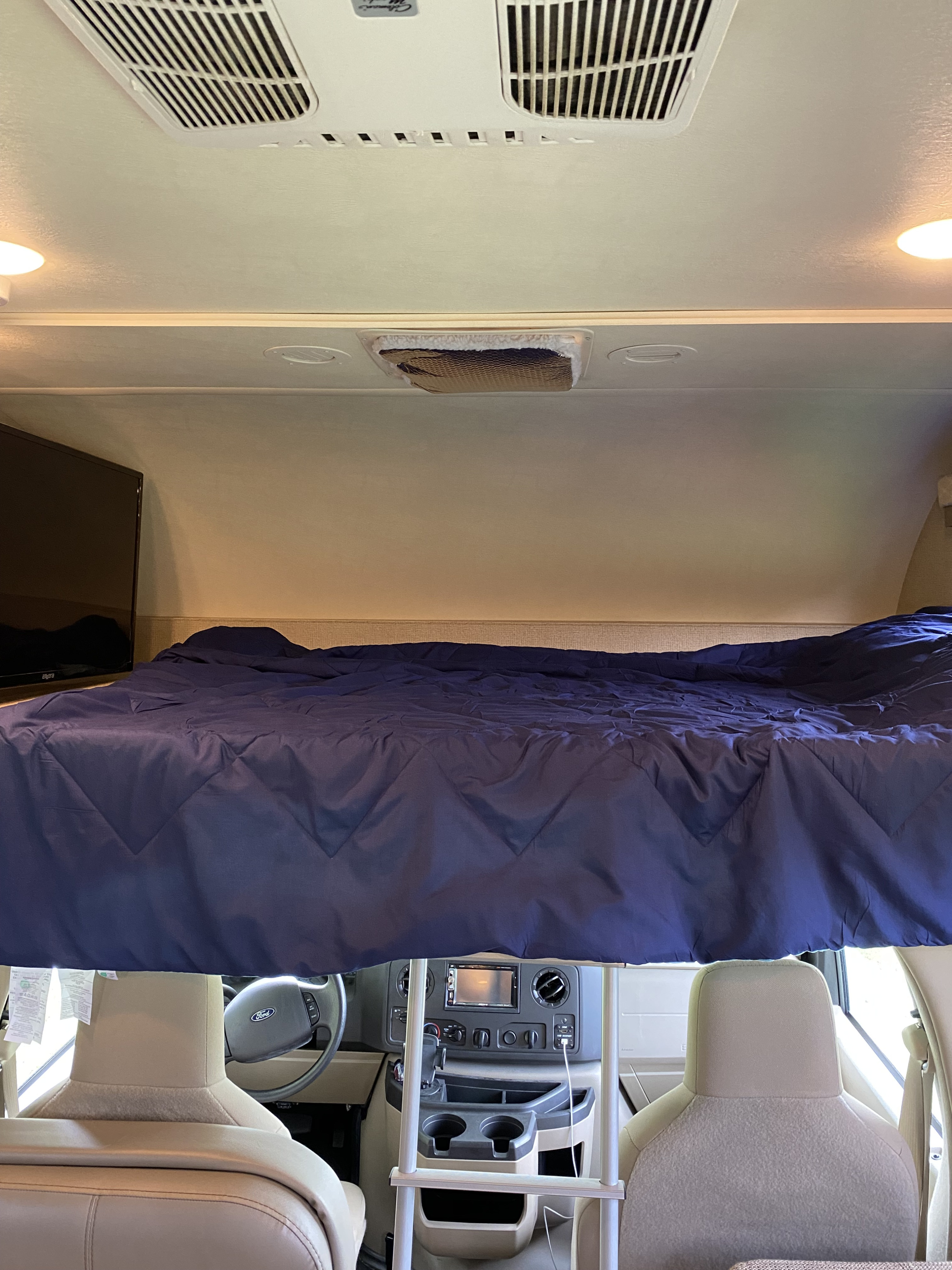 An RV rental interior shows the overhead bunk with blue bedding, a TV, and the Ford cab dashboard and steering wheel.. Thor Freedom Elite 2019