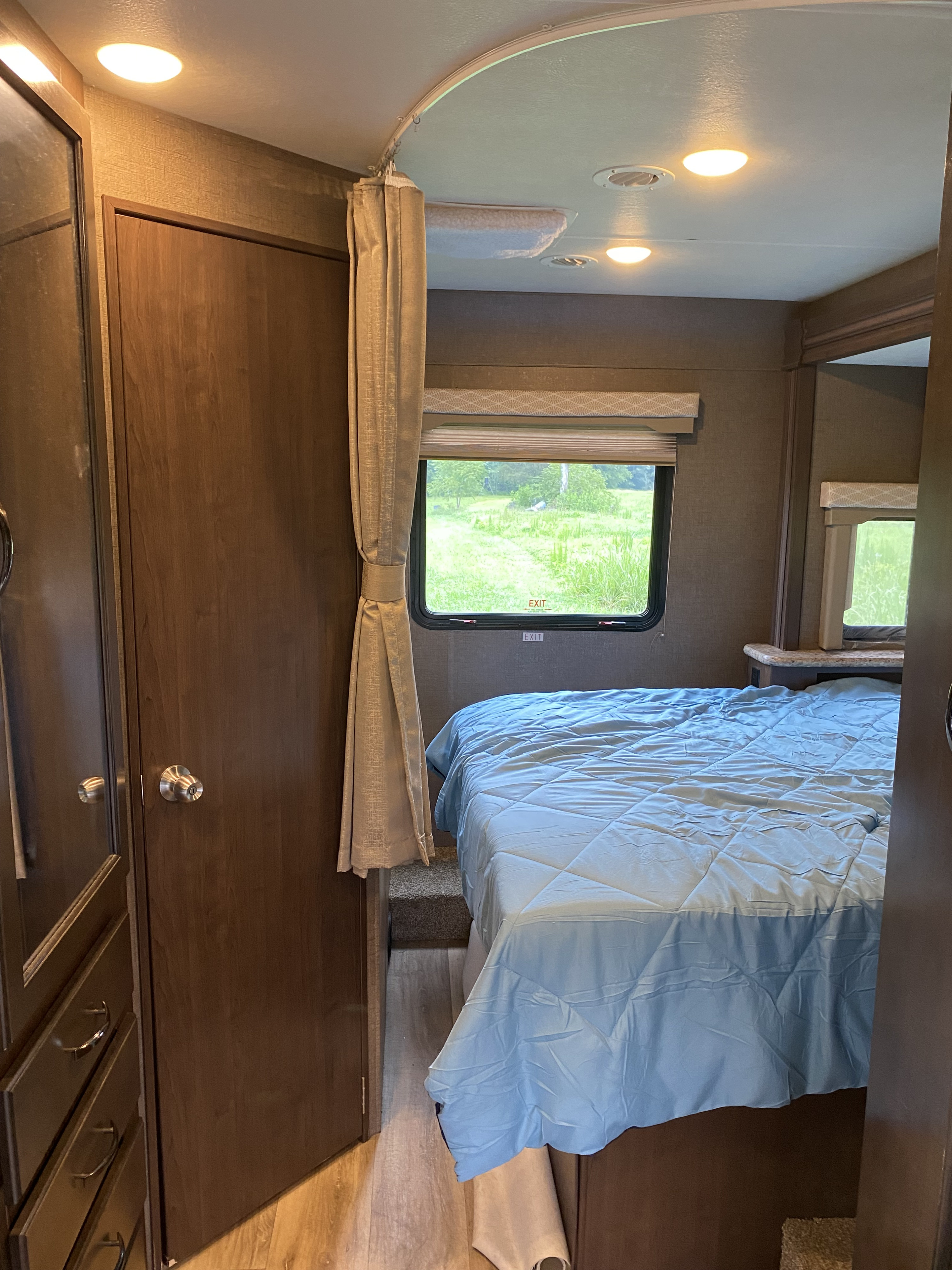 Cozy RV rental bedroom features a blue comforter, built-in storage, and a window showcasing a green outdoor view.. Thor Freedom Elite 2019