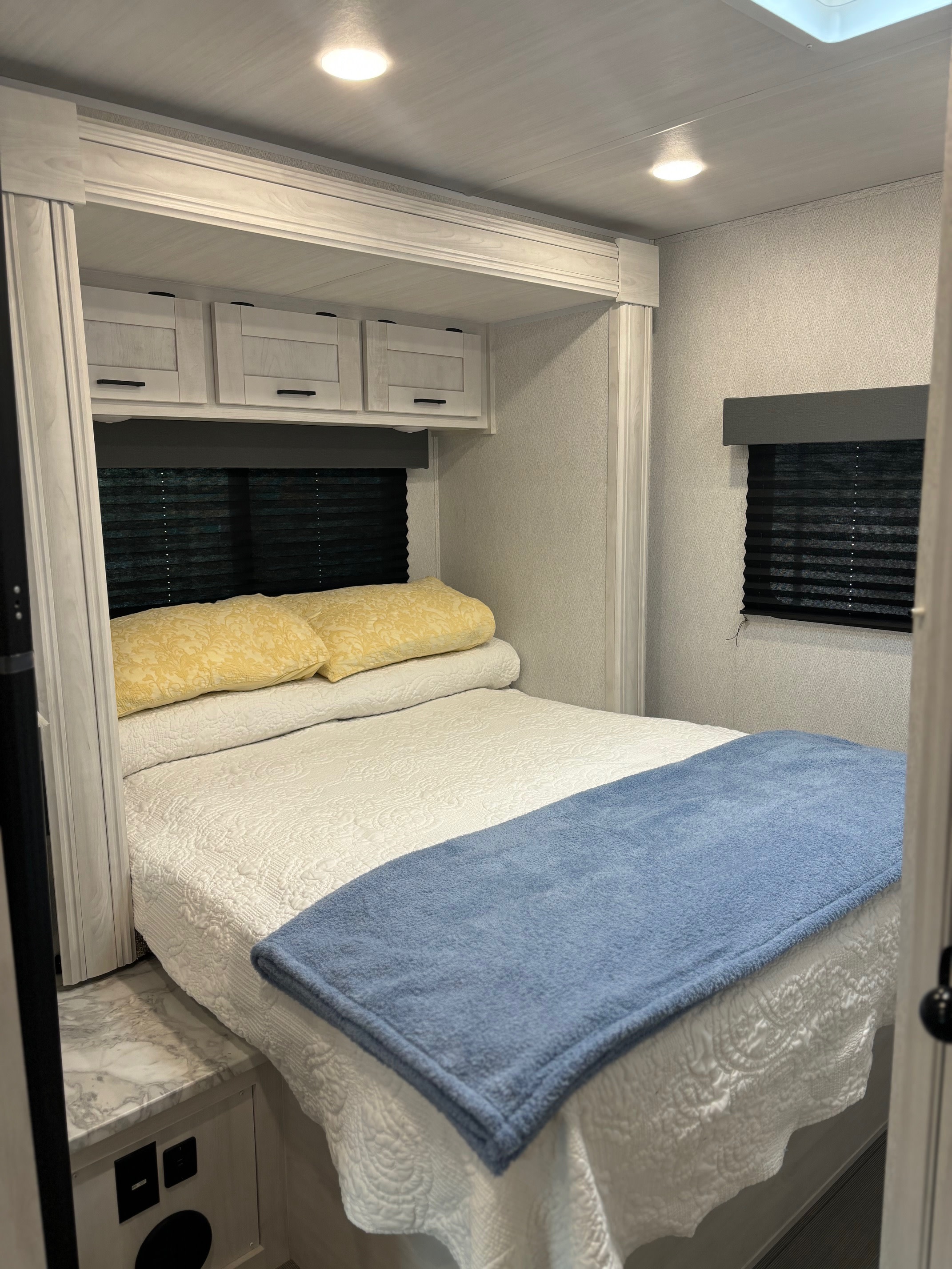 Bright, clean RV rental bedroom with a comfy bed, overhead cabinets, and windows with dark blinds.. Entrada East to West 2024