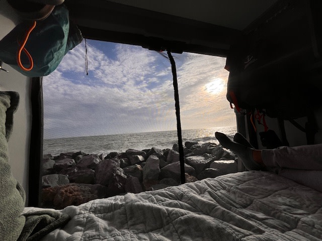 Cozy RV rental view: Sunrise/sunset over ocean, rocky shore. Feet propped on bed for ultimate relaxation.. Winnebago Solis 2023