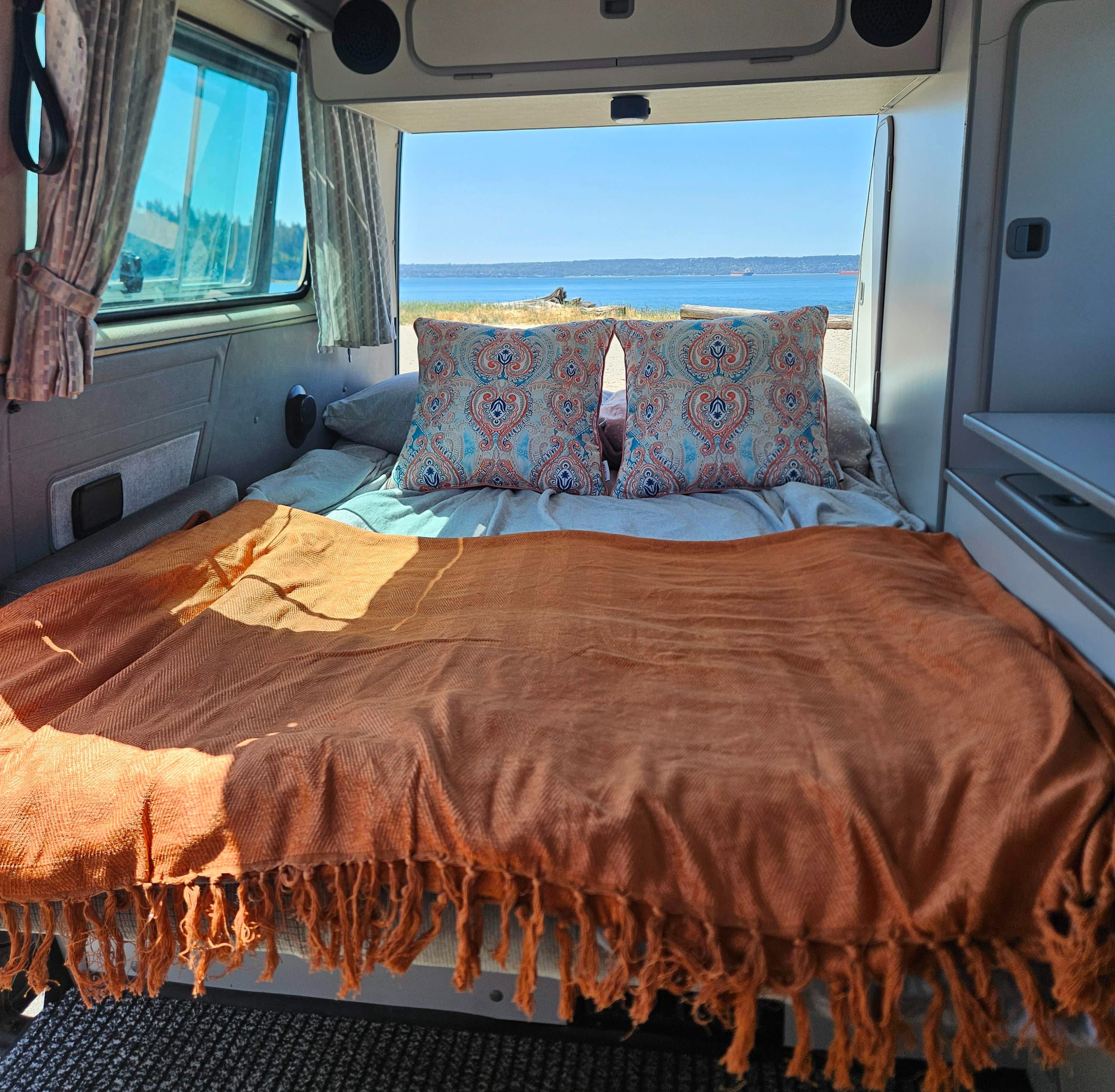 Inside an RV rental: a comfortable bed with patterned pillows & orange blanket, offering a scenic ocean view.. Westfalia T3 Westfalia 1987