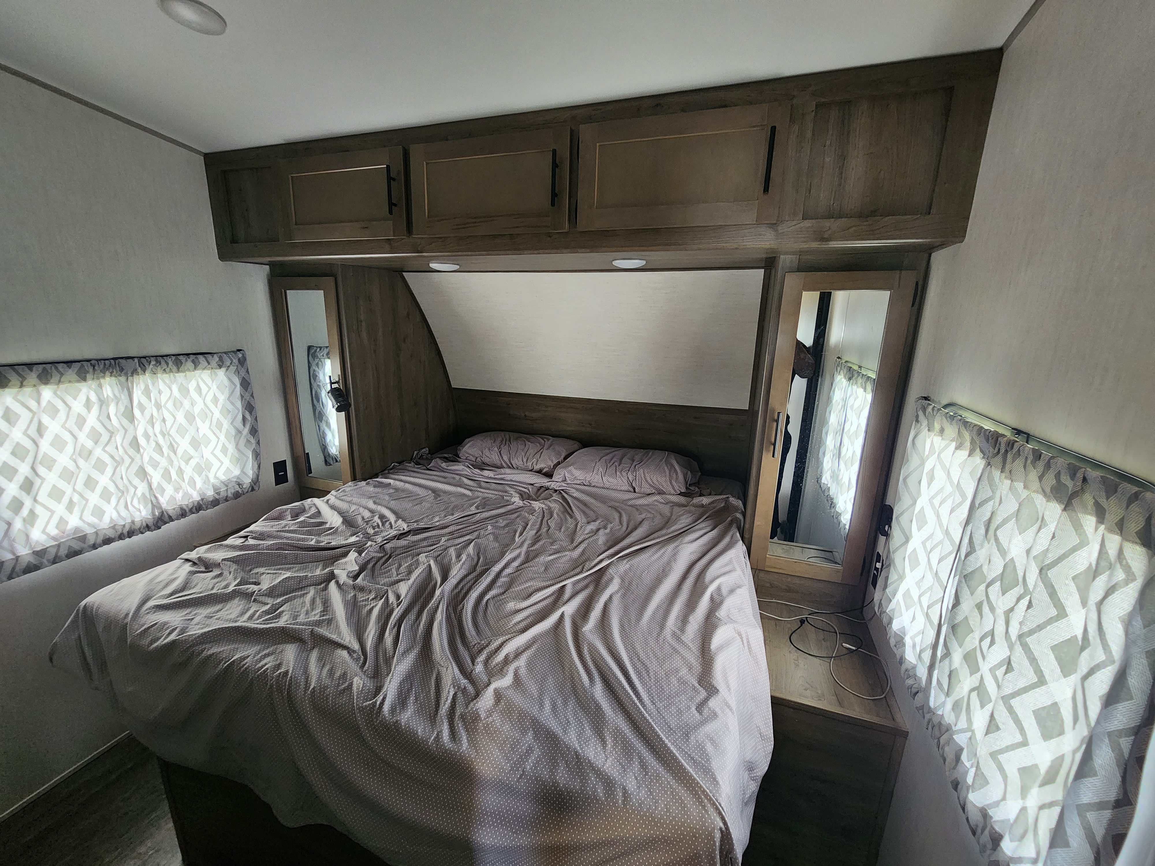 Cozy RV rental bedroom featuring a queen bed, overhead cabinets, and patterned window shades. Ideal for travel.. Forest River Wolf Pack 2023