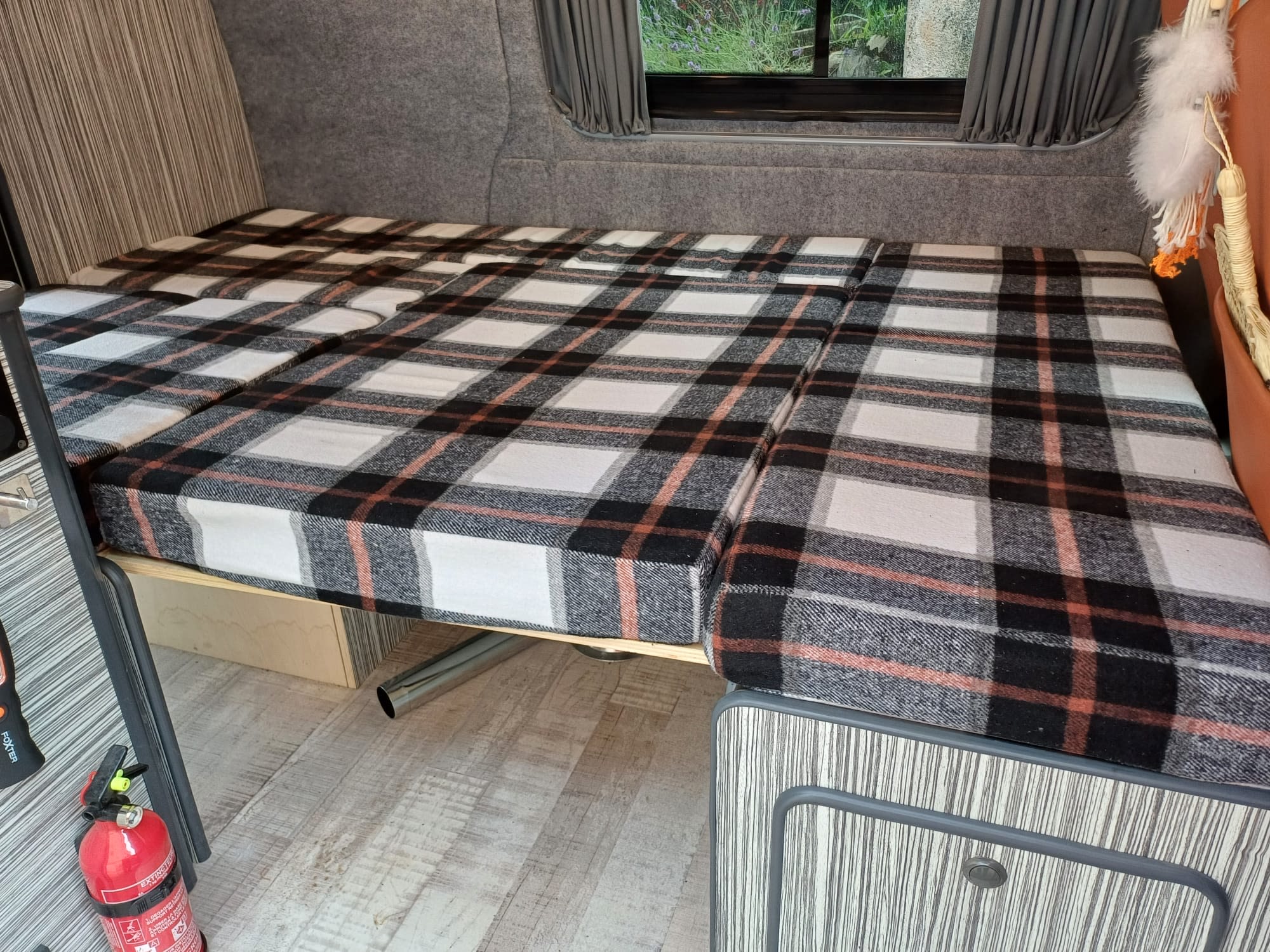 Comfortable plaid bed setup in an RV rental. Cozy camper interior with grey walls and wood-look floor.. Volkswagen T5 2012