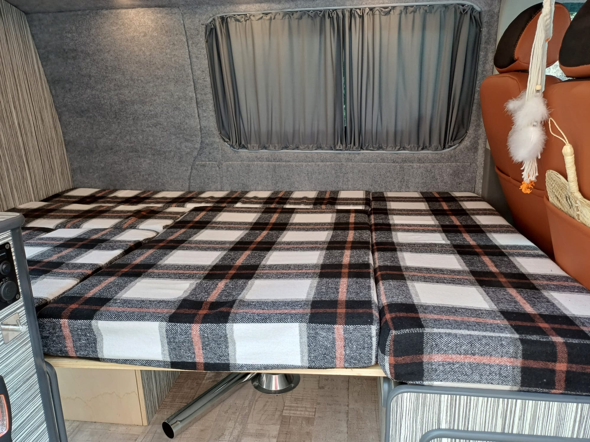 Cozy plaid mattress in an RV rental campervan interior, featuring grey walls, curtains, and tan seating.. Volkswagen T5 2012