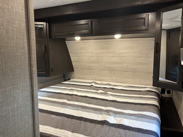 Cozy RV rental bedroom with a striped bed, overhead dark cabinets, and task lighting. Ideal for your next adventure.. Jayco Eagle 312 BHOK 2024