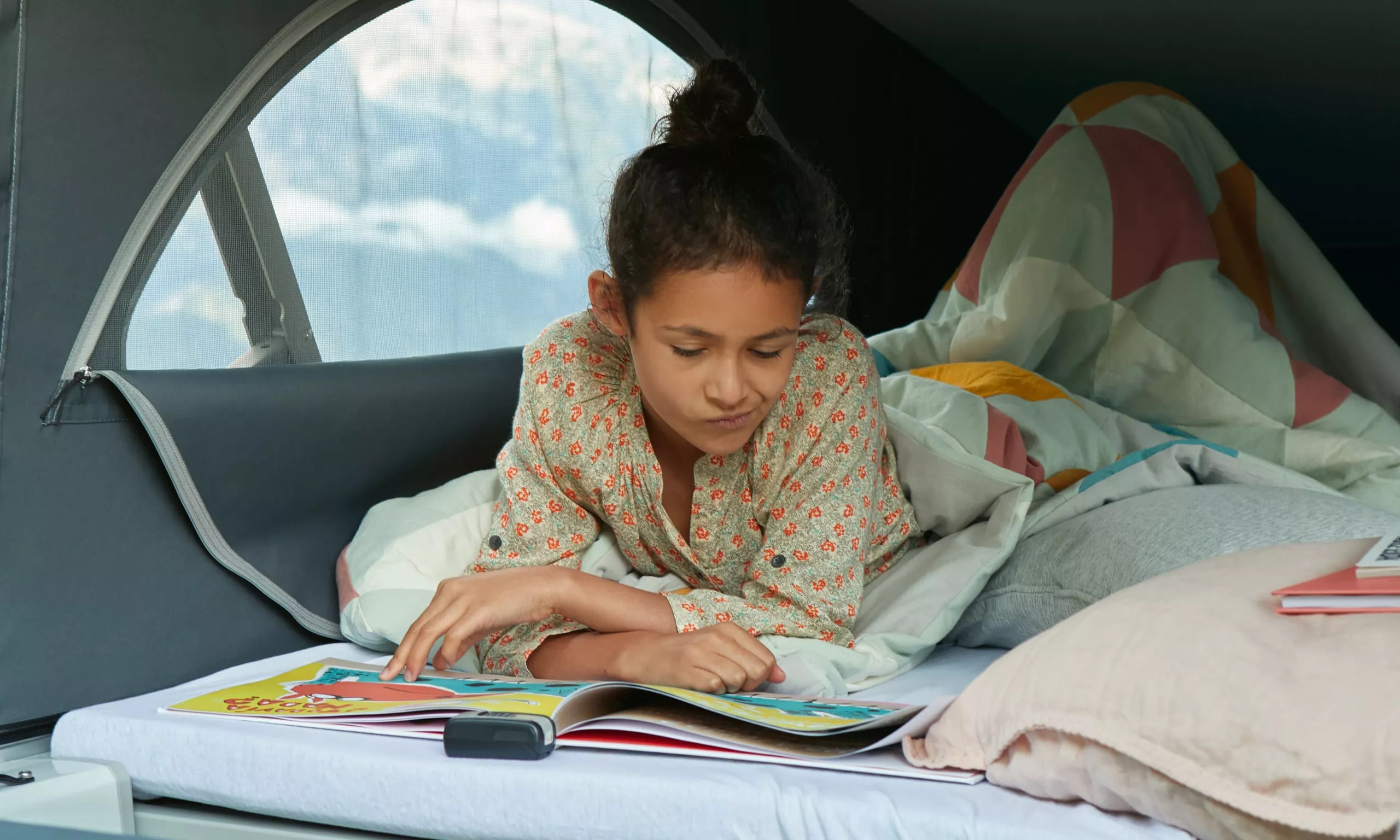 An RV rental offers cozy comfort. Young girl enjoys reading a book on a camper bed, window showing cloudy sky.. Volkswagen California T6.1 4MO 2024
