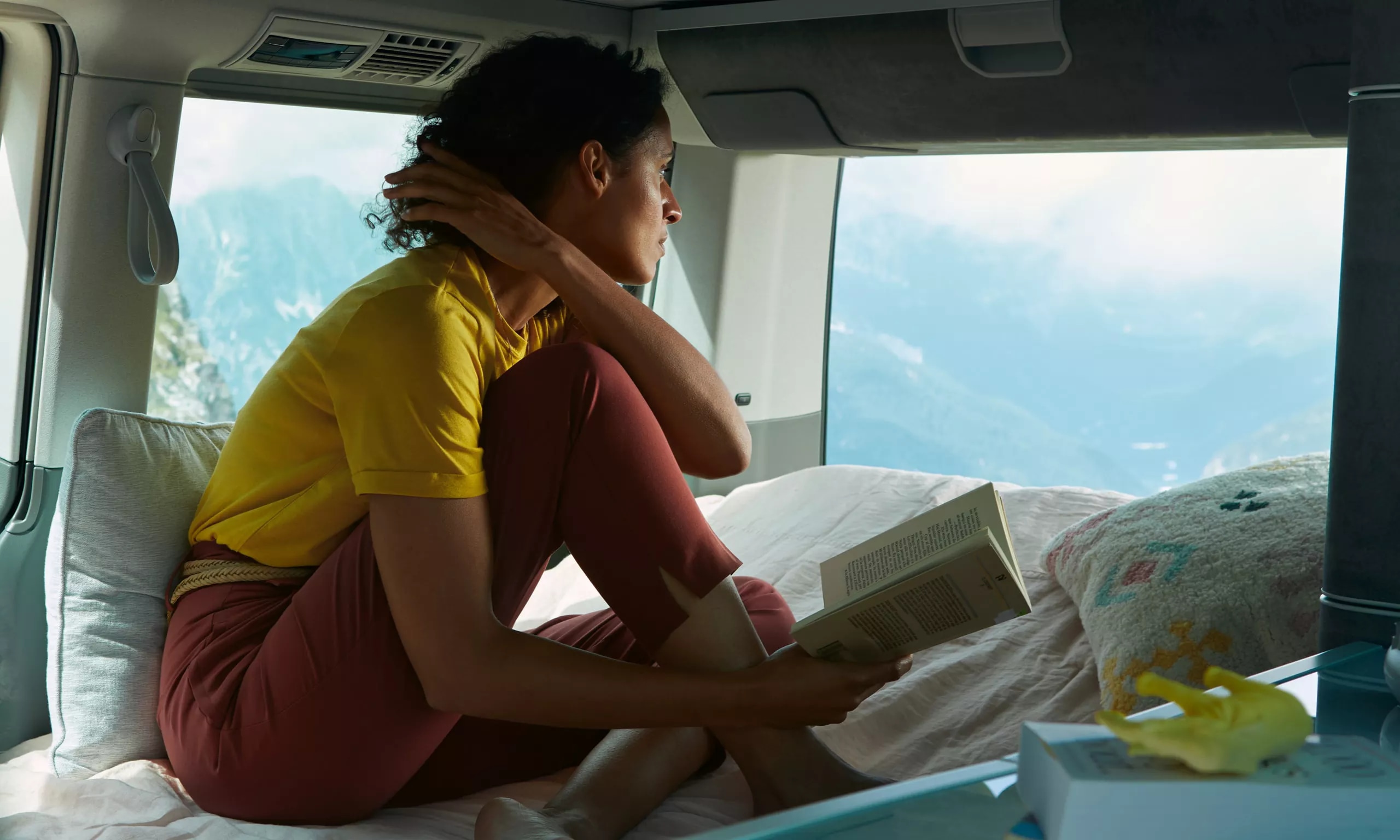 A woman enjoys her RV rental, reading a book in a cozy interior with stunning mountain views.. Volkswagen California T6.1 4MO 2024