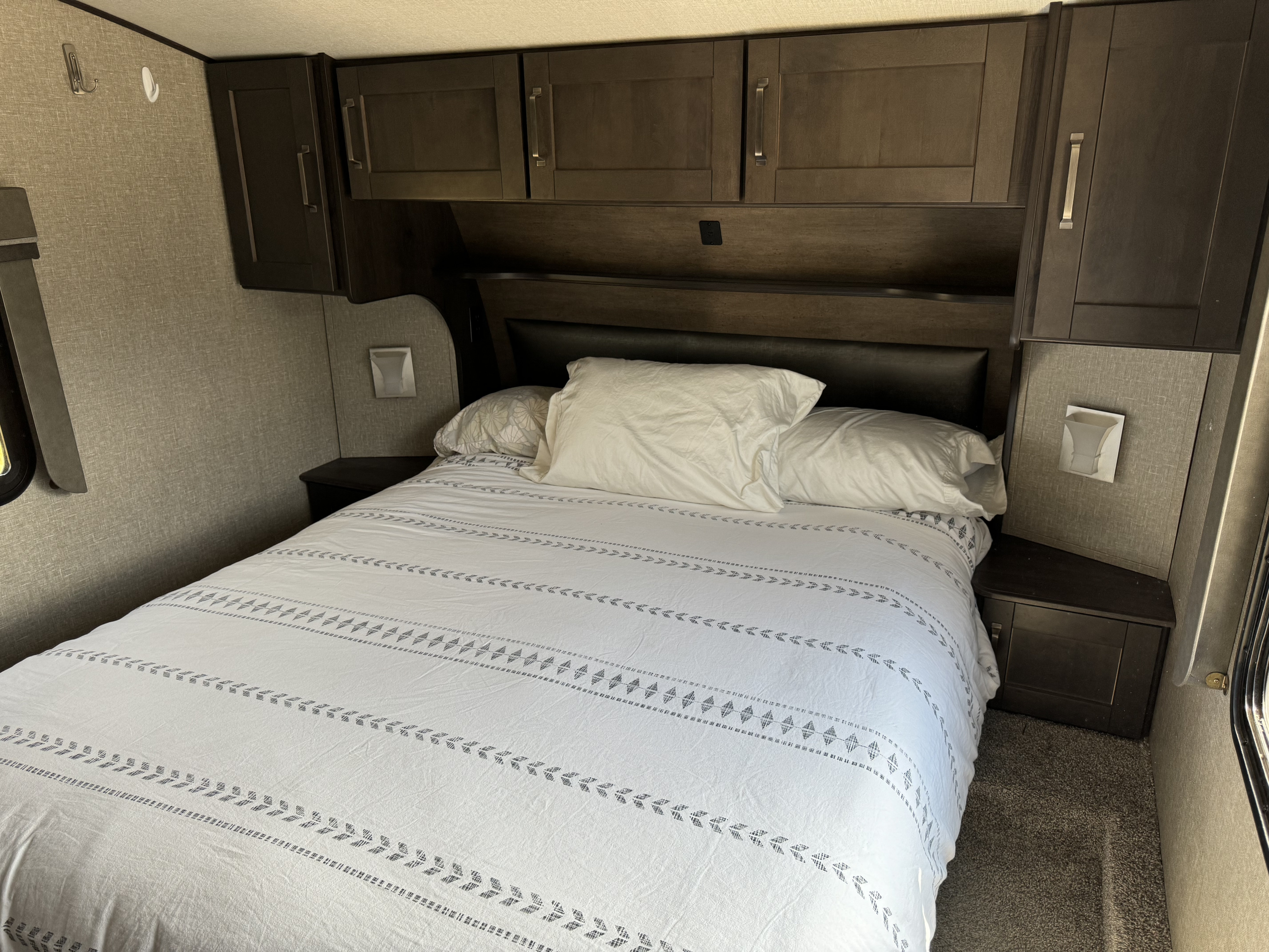 This **RV rental** bedroom offers a comfortable bed with white bedding, dark cabinets, and convenient nightstands.. Grand Design Reflection 2021