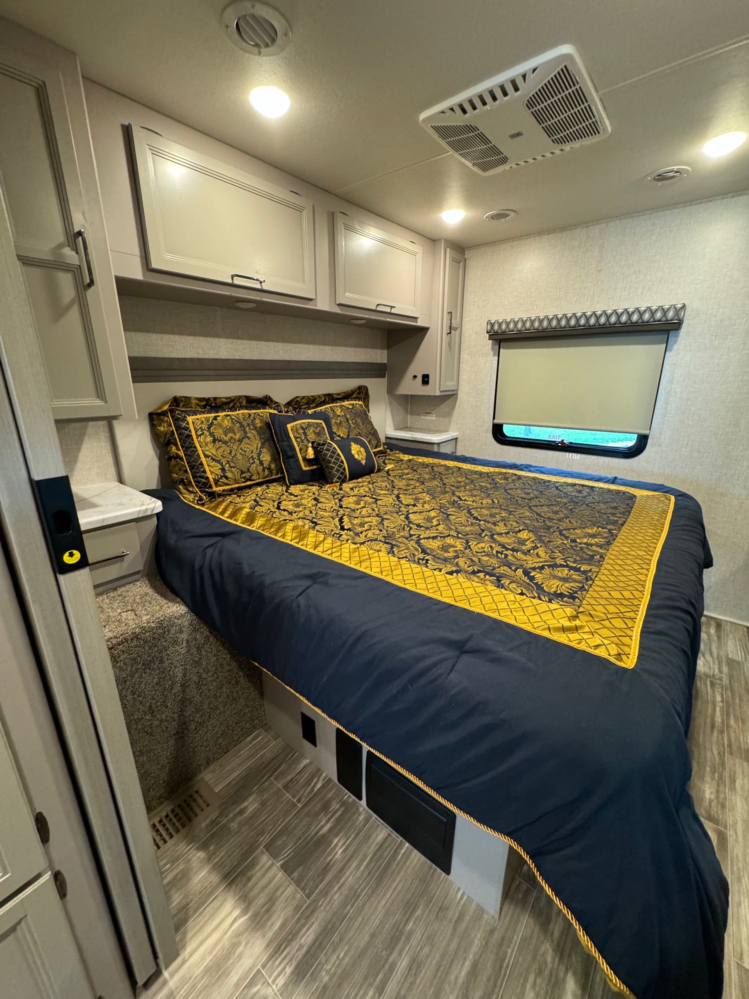 Cozy RV rental bedroom featuring elegant blue and gold bedding, overhead storage, and a window with a blind.. Thor Hurricane 2023