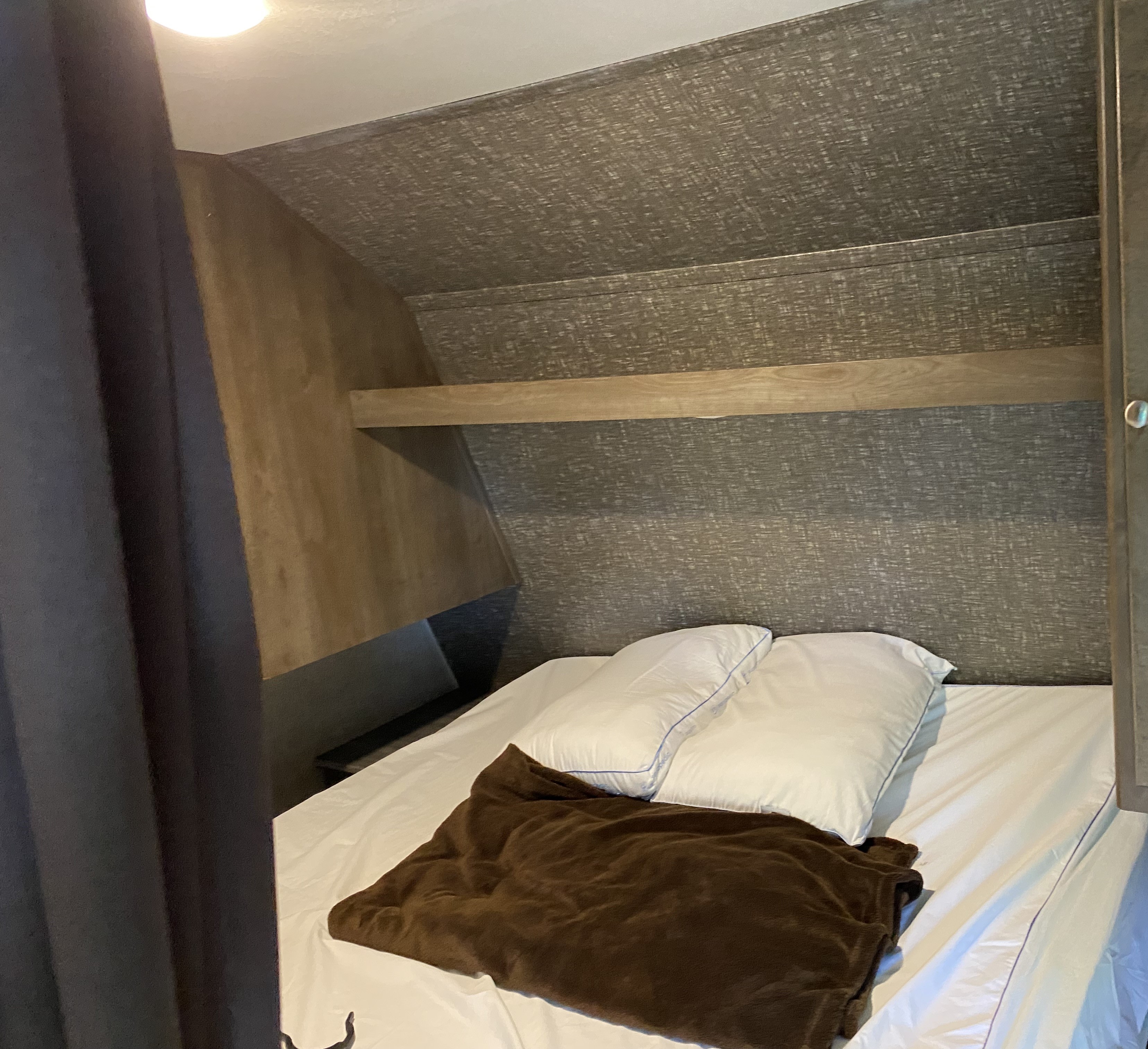 Cozy RV rental bedroom with white linen bed, pillows, brown blanket, textured walls, and a wooden shelf.. Keystone RV Coleman Lantern edition 295QB 2019