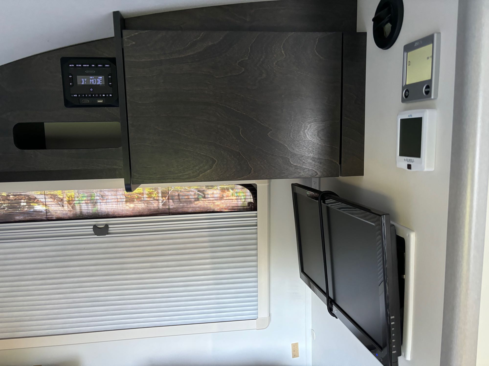 Modern RV rental interior: Wall-mounted TV, multi-media stereo ('BT MODE'), climate controls, and a window with blinds.. nuCamp T@G 2022