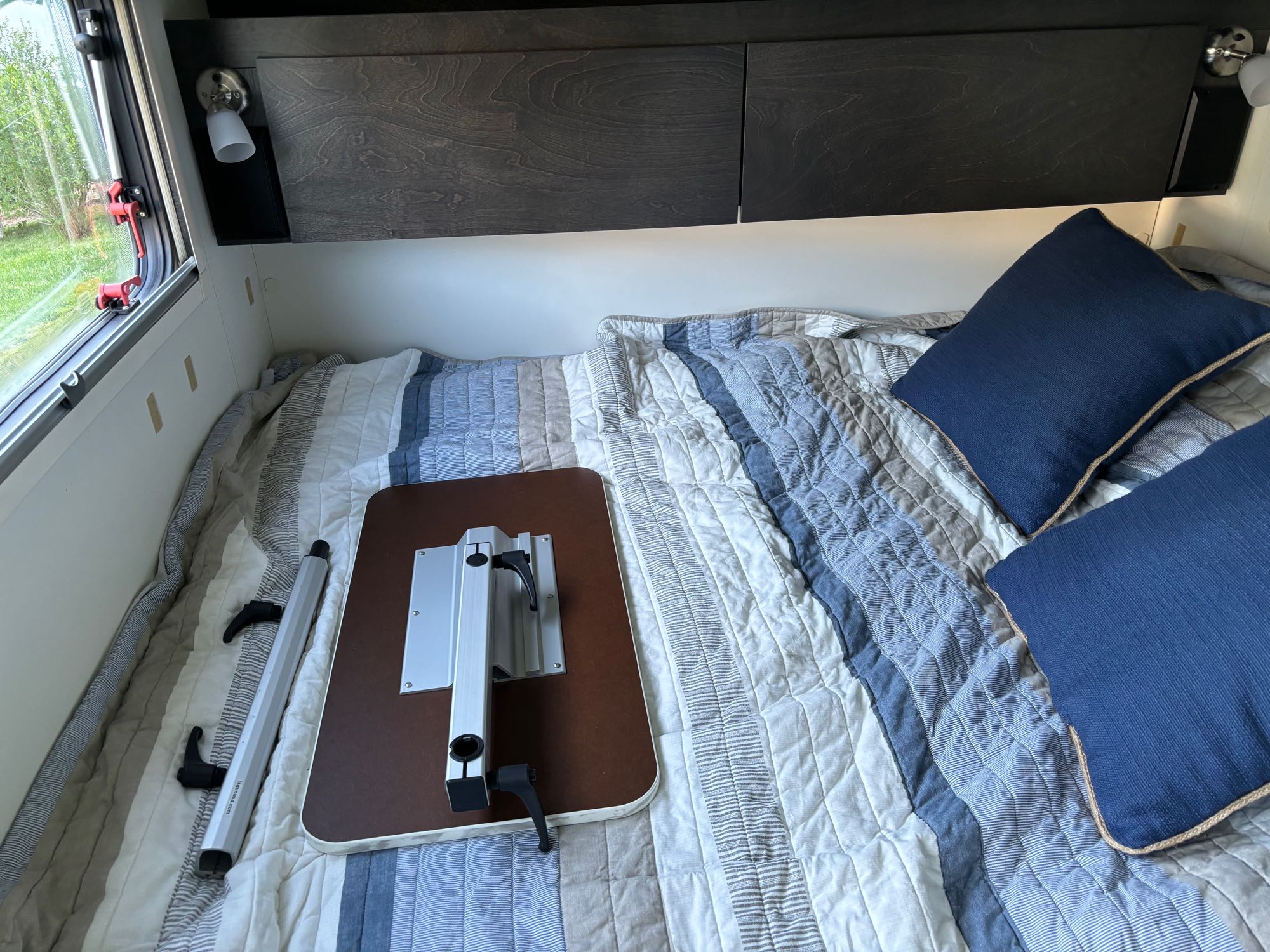 This RV rental's bedroom shows a striped bed, blue pillows, overhead cabinets, and a table disassembled for travel.. nuCamp T@G 2022