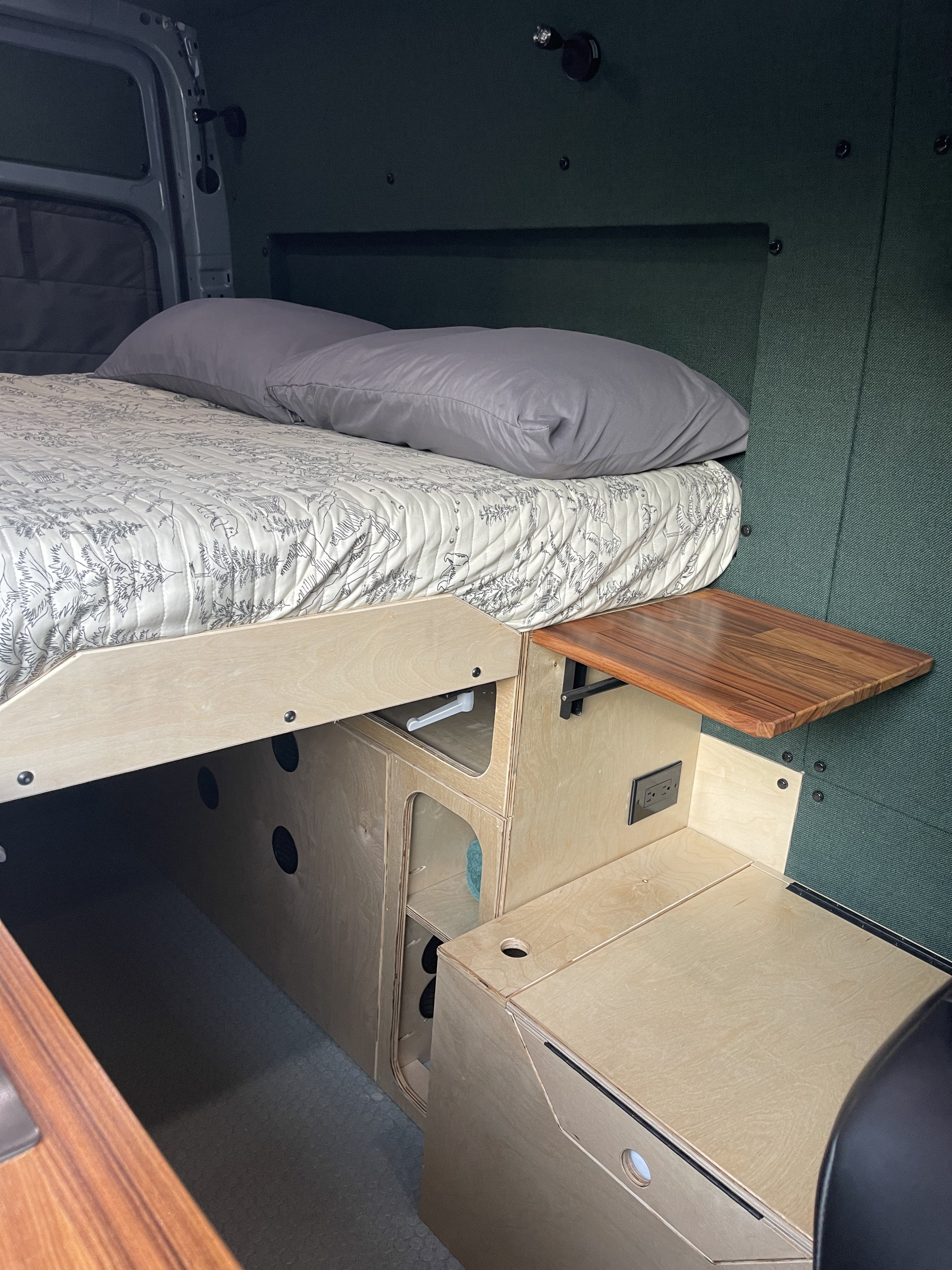 Inside a cozy RV rental: a comfortable bed with a patterned quilt, integrated storage, and a functional side table.. Mercedes Sprinter 2021