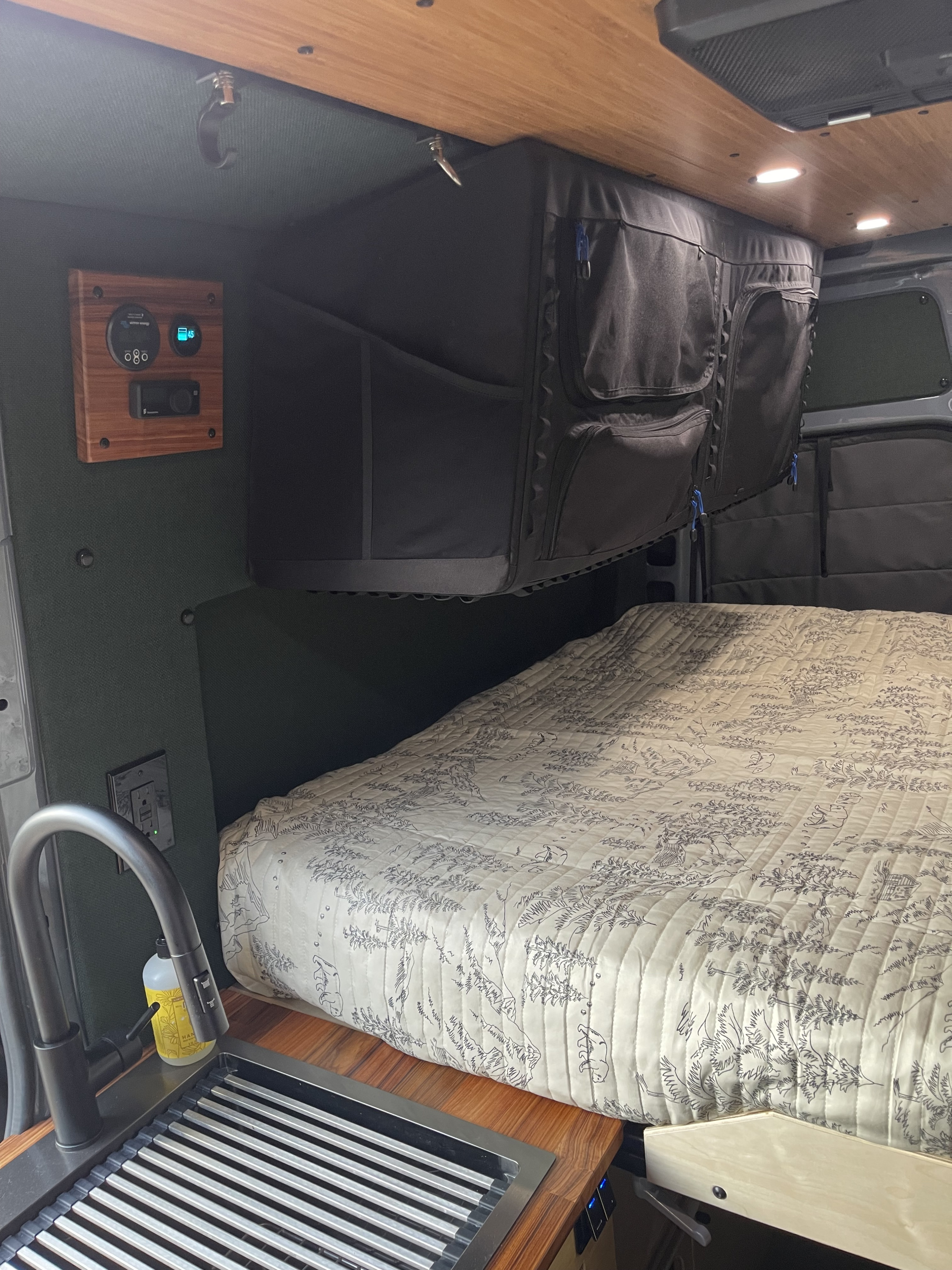 Cozy RV rental interior: bed with forest-patterned quilt, kitchen sink, wood accents, smart controls, and overhead storage.. Mercedes Sprinter 2021