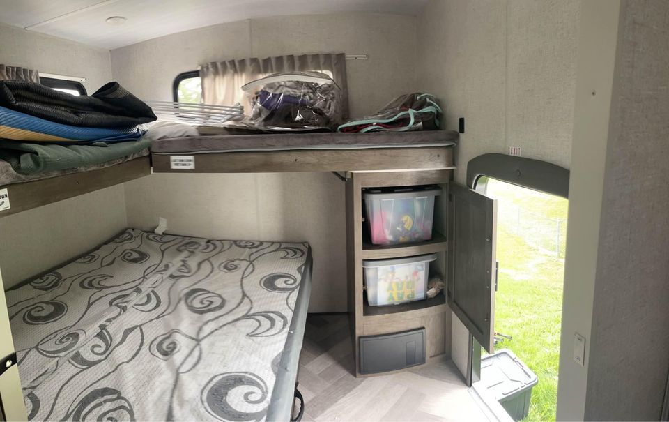 RV rental interior with stacked bunk beds, built-in storage shelves holding clear bins, and an open exit door revealing grass outside.. Forest River Wildwood 2021