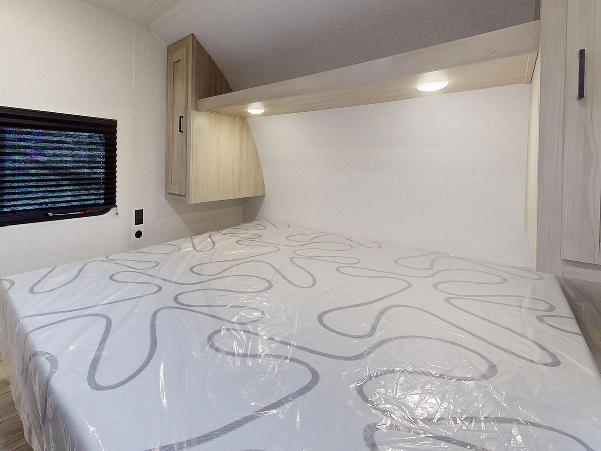 RV rental bedroom featuring a bed with a plastic-covered mattress, overhead cabinets, and a window with blinds.. East to West Silver Lake 3250QB 2025