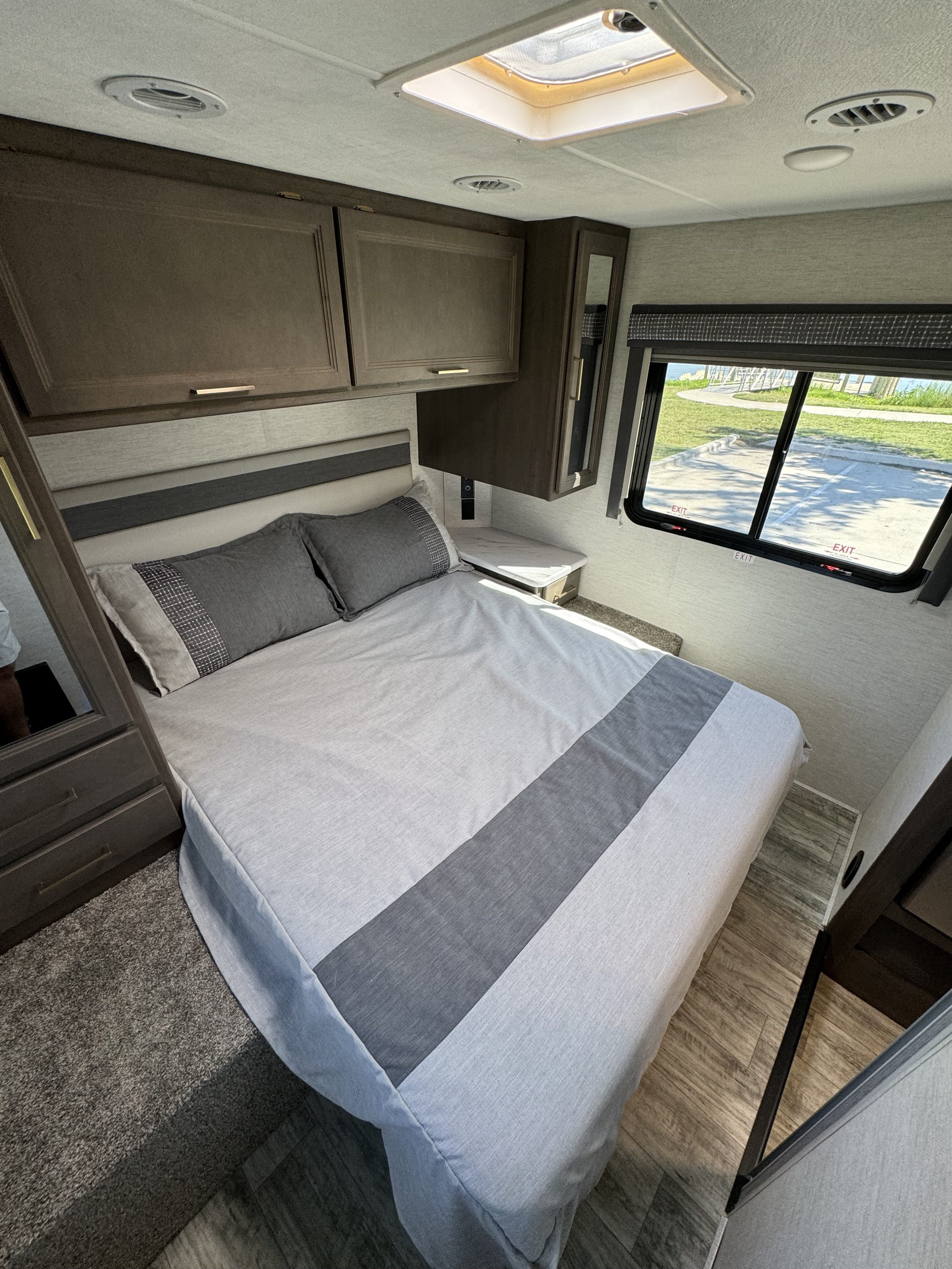 Cozy RV rental bedroom featuring a queen bed with gray bedding, overhead cabinets, and a scenic window view.. Thor Chateau 2024