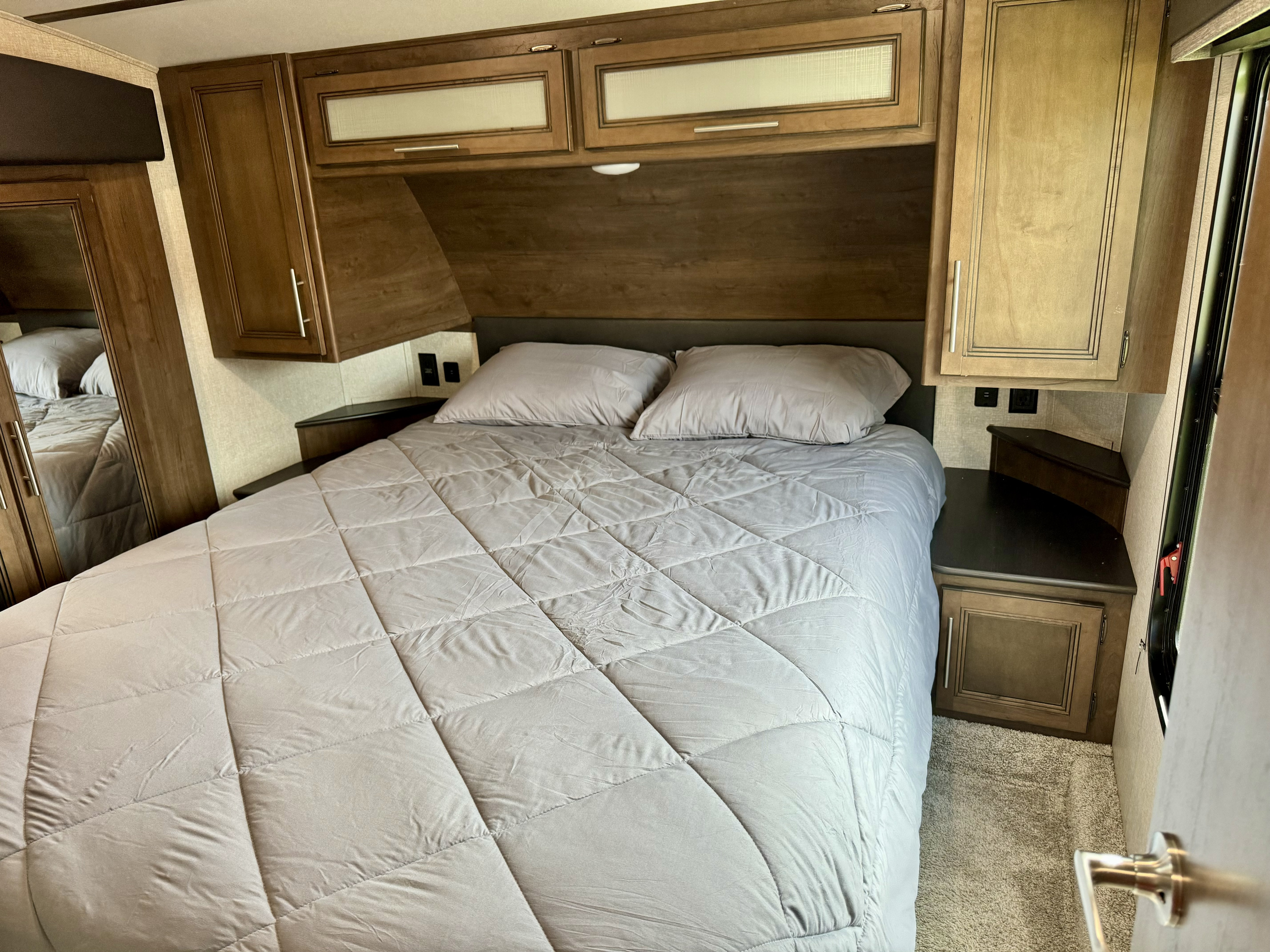 RV rental bedroom featuring a cozy bed with gray bedding & pillows, overhead and side wooden cabinetry, & carpeted floor.. Keystone RV Cougar 2020