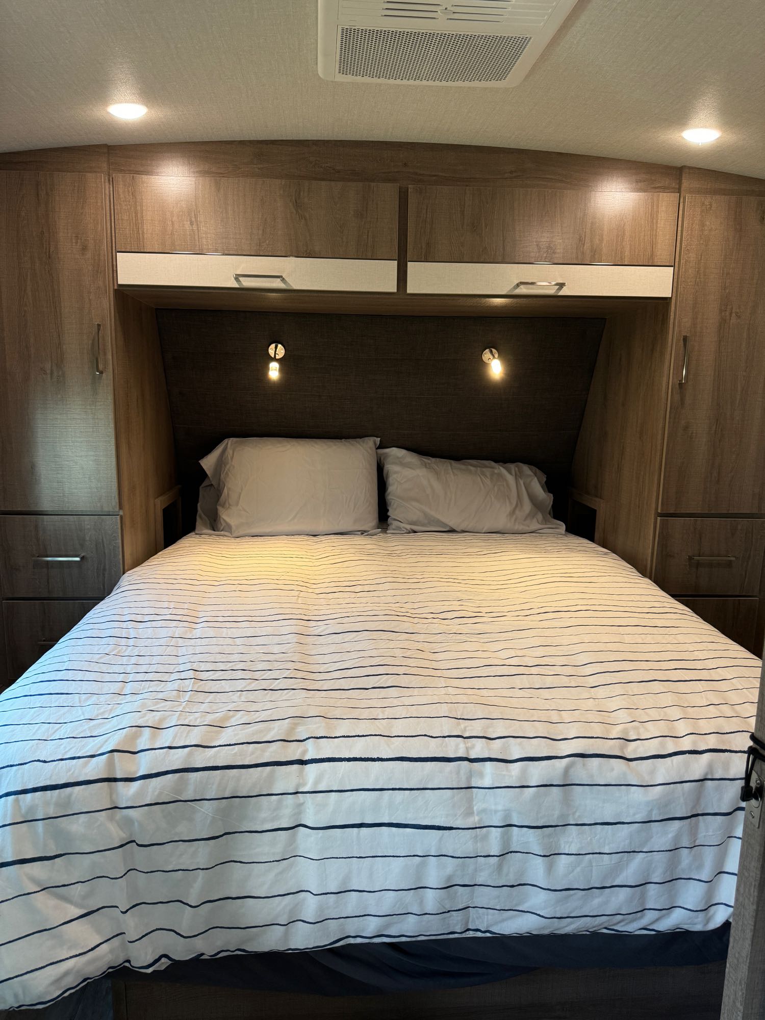 Cozy RV rental bedroom with striped bedding, two pillows, overhead cabinets, and wall-mounted lights.. Grand Design Imagine 2022