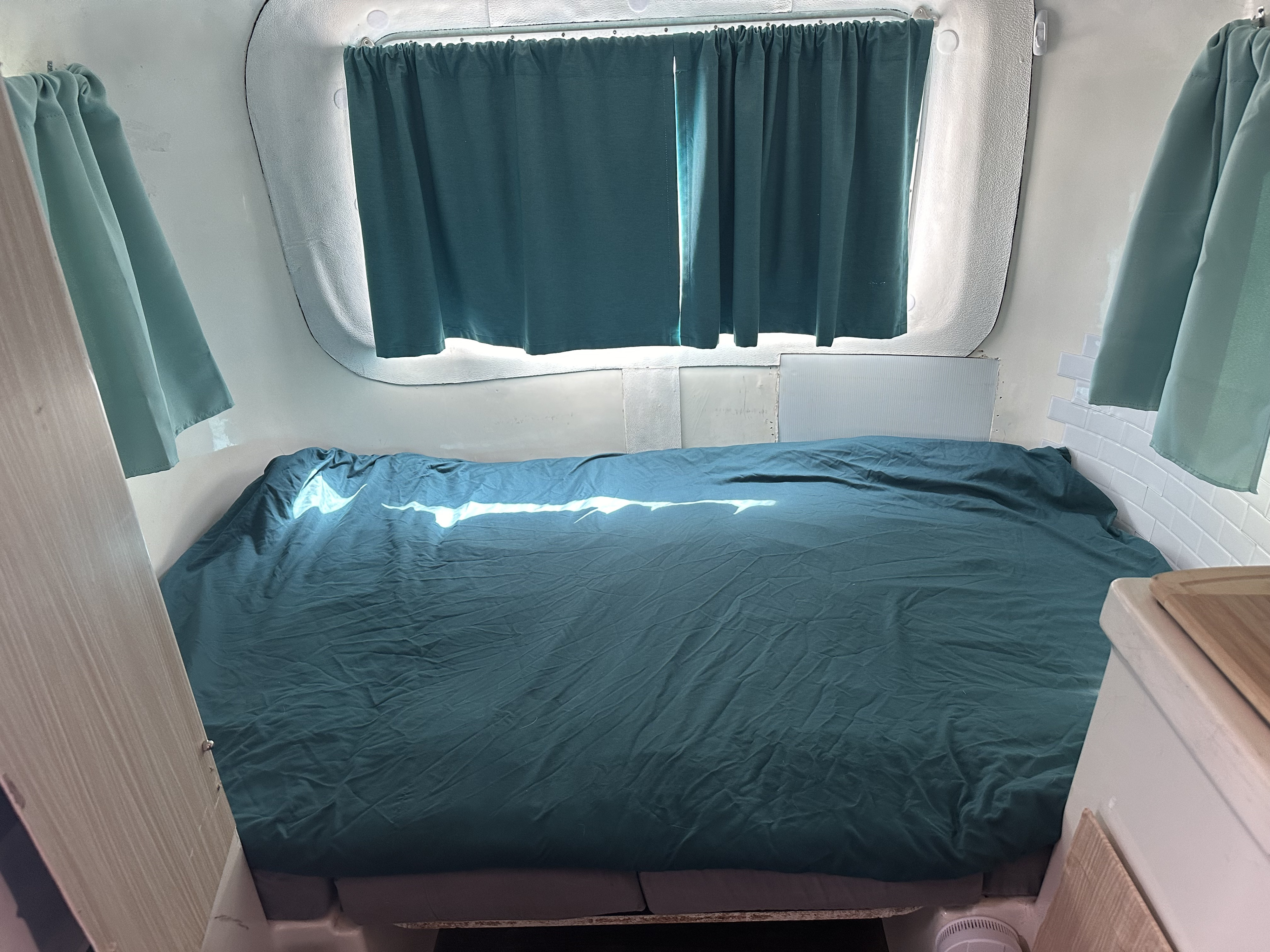 Comfortable sleeping area inside an RV rental, featuring a bed with teal covers and matching curtains. Sunlight illuminates the space.. Burro 10 foot 1978