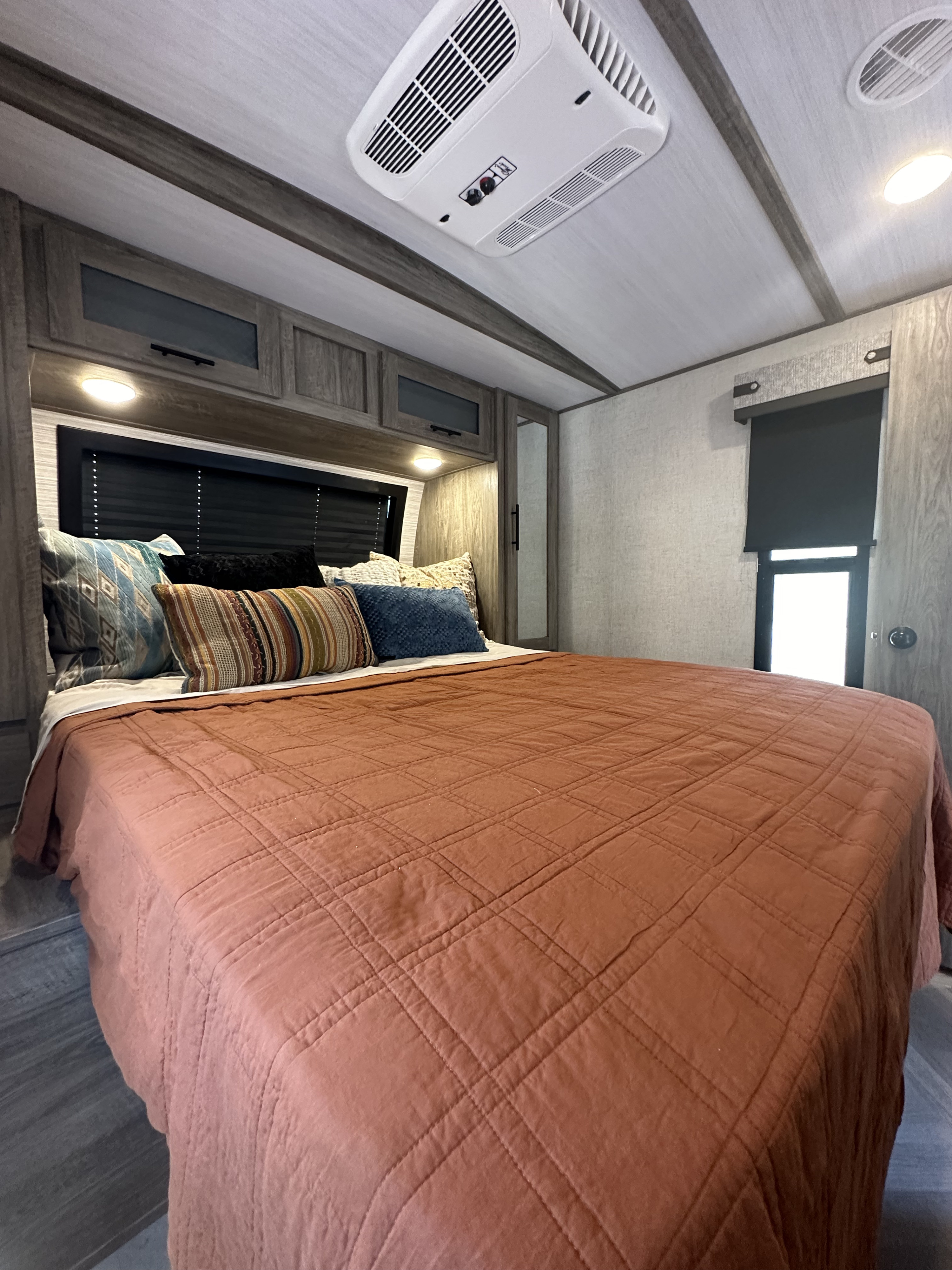 Modern RV rental bedroom with a large bed, rust-colored quilt, overhead cabinets, an AC unit, and dark roller shades.. Forest River Salem Hemisphere 2021