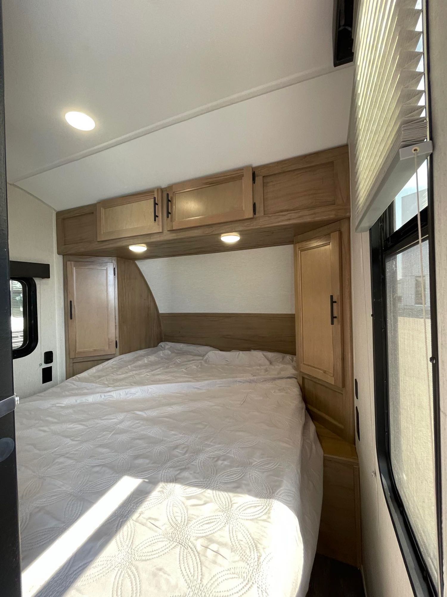 RV rental bedroom with a large bed, white quilt, overhead cabinets, side wardrobes, and sunny window.. Forest River Cherokee Wolf Pup 2023