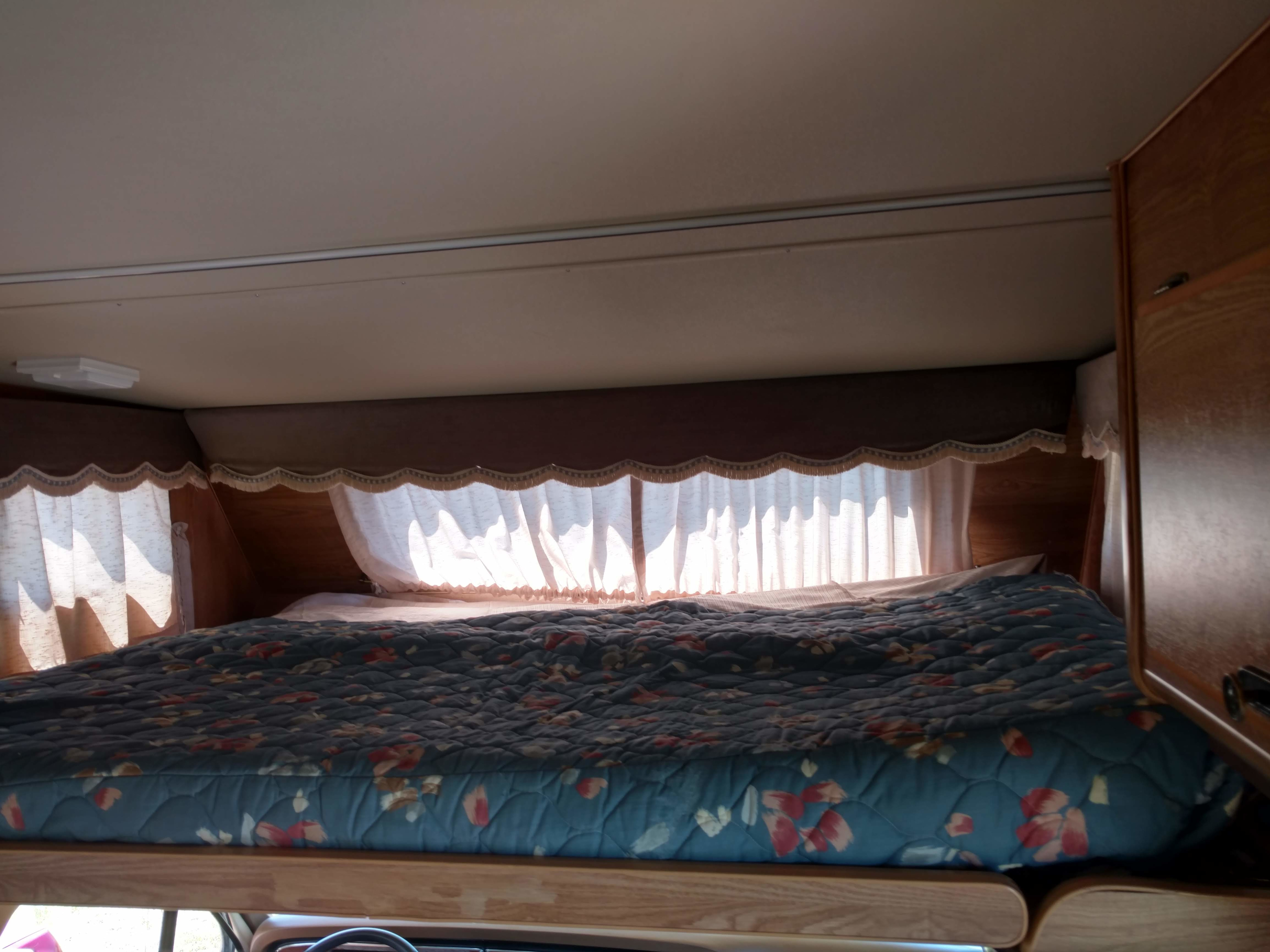 Cozy RV rental bed with floral quilt and bright windows, perfect for travel accommodations.. Ford E350 Tioga Arrow 1988