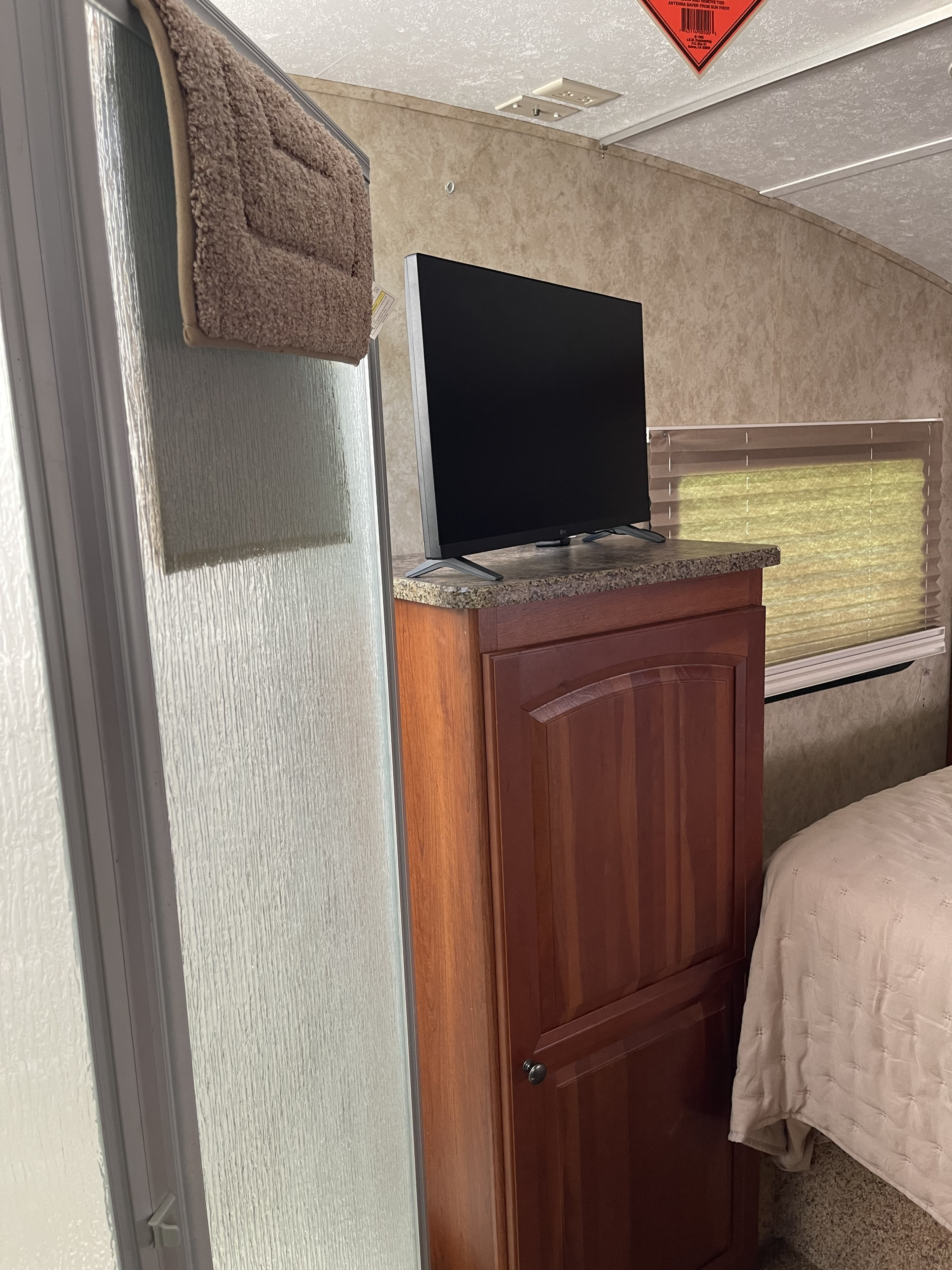 RV rental interior: TV on a wooden cabinet, textured shower door with hanging bath mat, window blinds, and part of a bed.. Keystone RV Cougar 2010