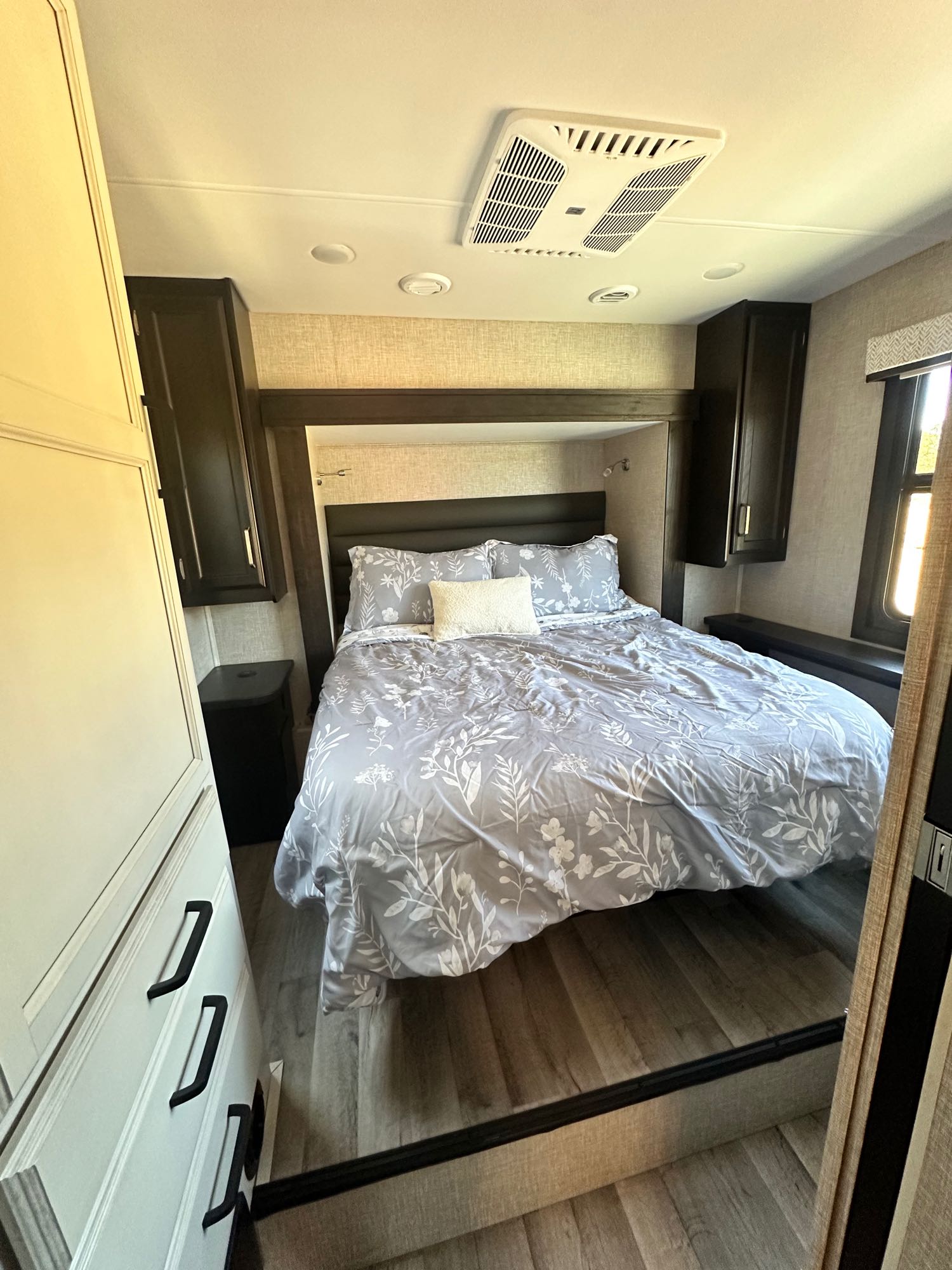 RV rental bedroom: cozy bed with grey floral bedding, ceiling AC unit, and built-in storage.. Jayco Greyhawk 2023