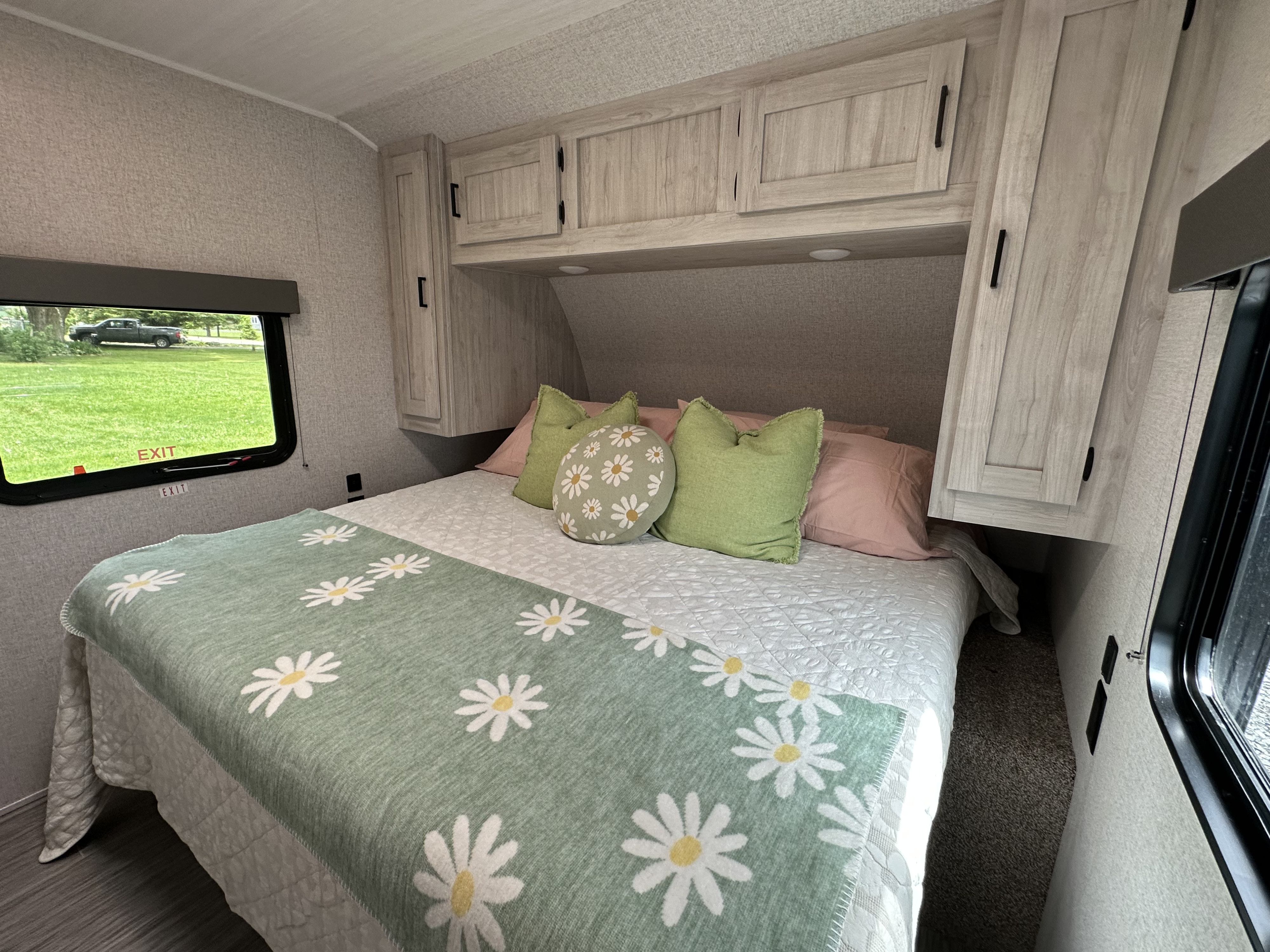 Cozy RV rental bedroom with a comfy bed, daisy-patterned blanket, overhead storage, and an outdoor view through the window.. East to West Silver Lake 2023