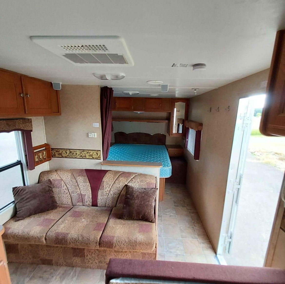 Cozy RV rental interior featuring a patterned sofa lounge, connected to a bedroom with a blue mattress, under wood cabinets.. Dutchmen Lite 2007