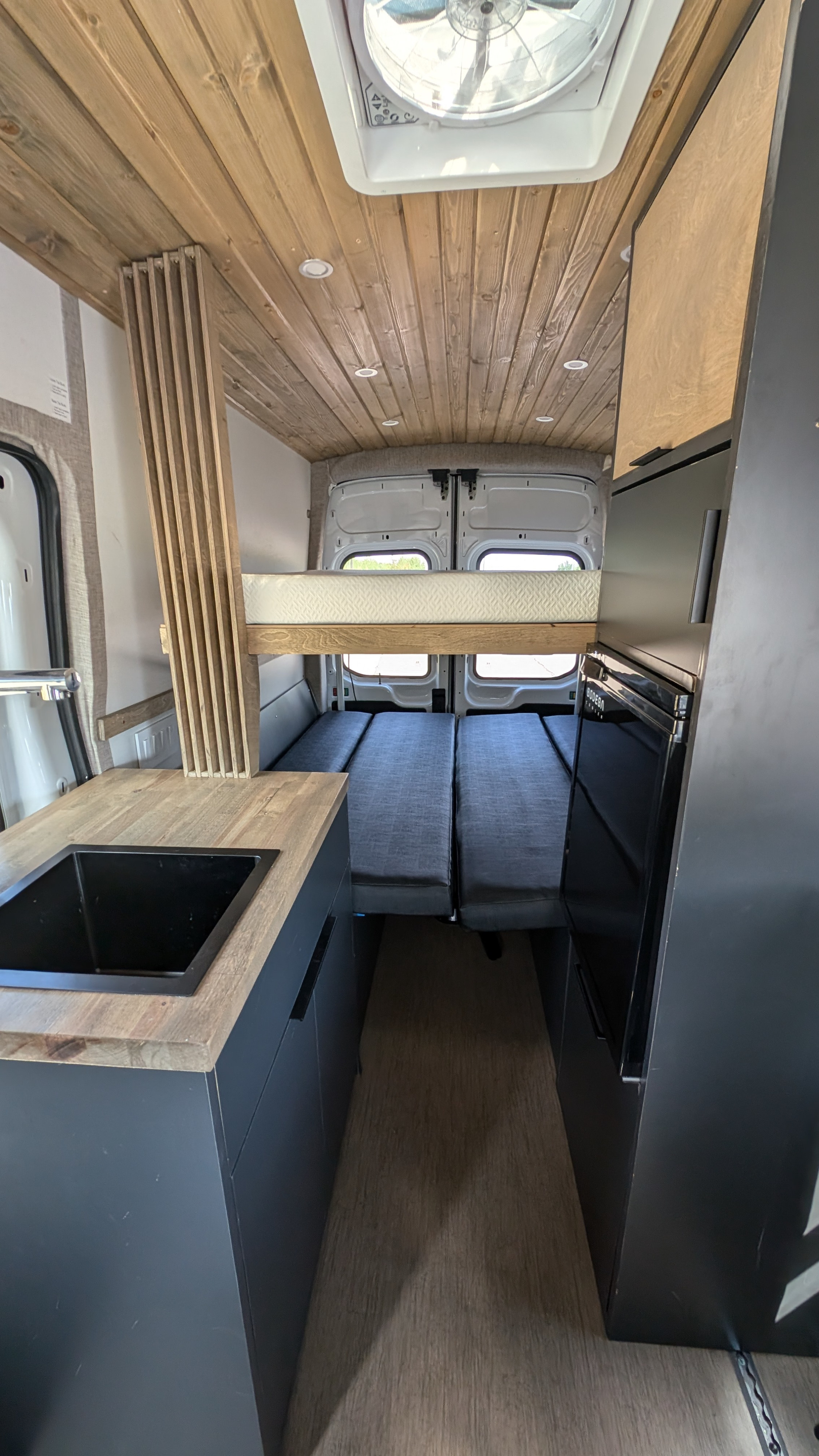 Compact RV rental interior: custom kitchen with sink, wood-paneled ceiling, and a versatile bed/seating area.. Ford Van 2024