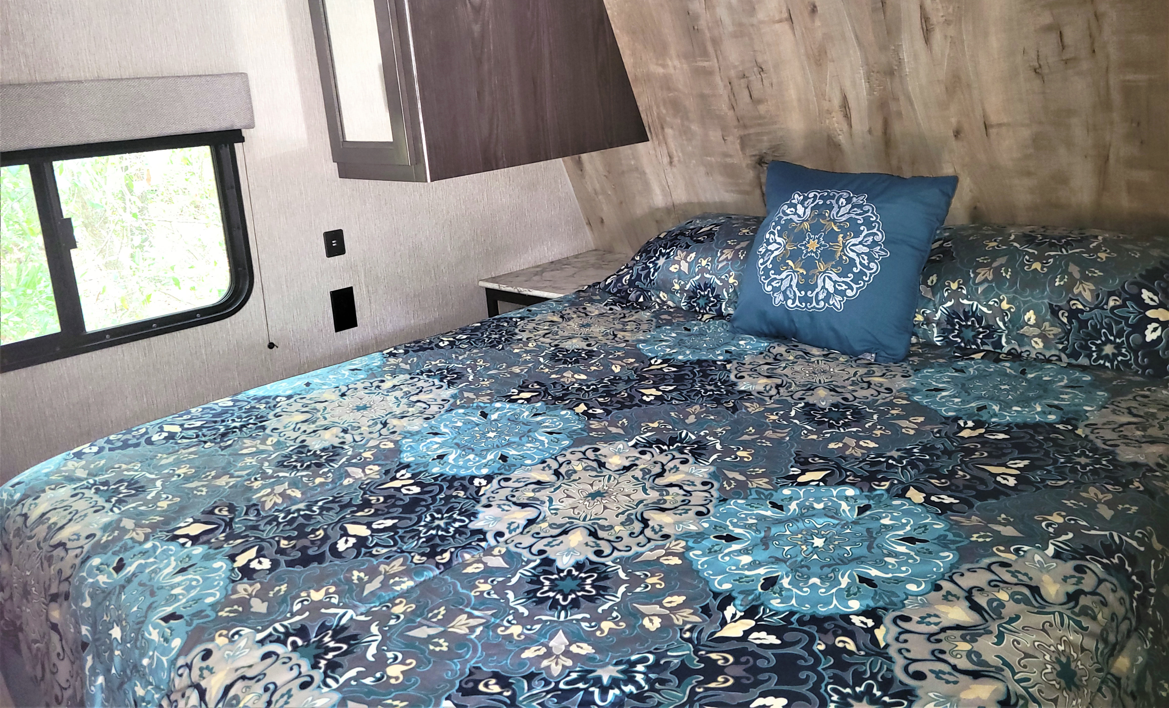 Inside a comfortable RV rental bedroom, featuring a blue patterned bed, window view, and overhead cabinet.. Guide Guide Trailer 2020