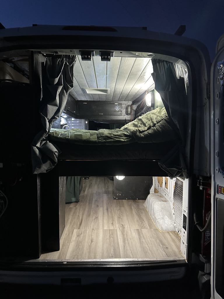 Inside this RV rental camper van: A comfy bed over a roomy living area with wood floors, perfect for camping.. Ford Transit 2017
